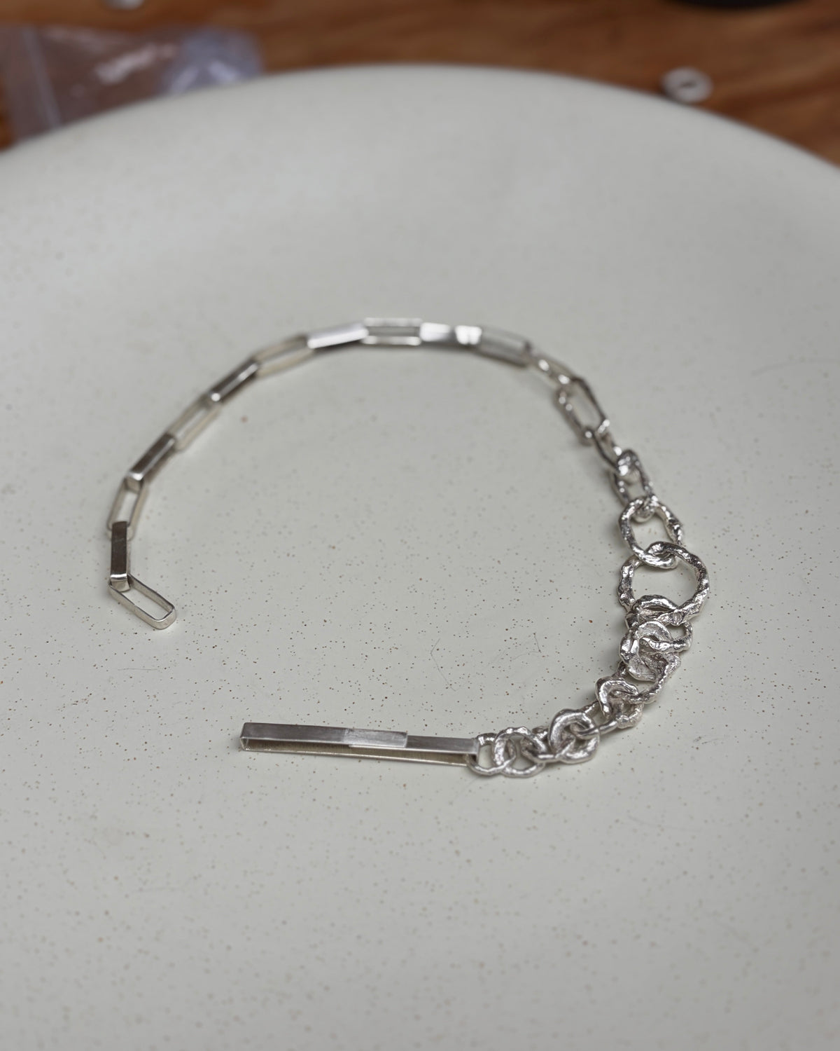 A silver-toned metal bracelet with a combination of flat links, chain links, and a rectangular bar clasp, resting on a light speckled surface.