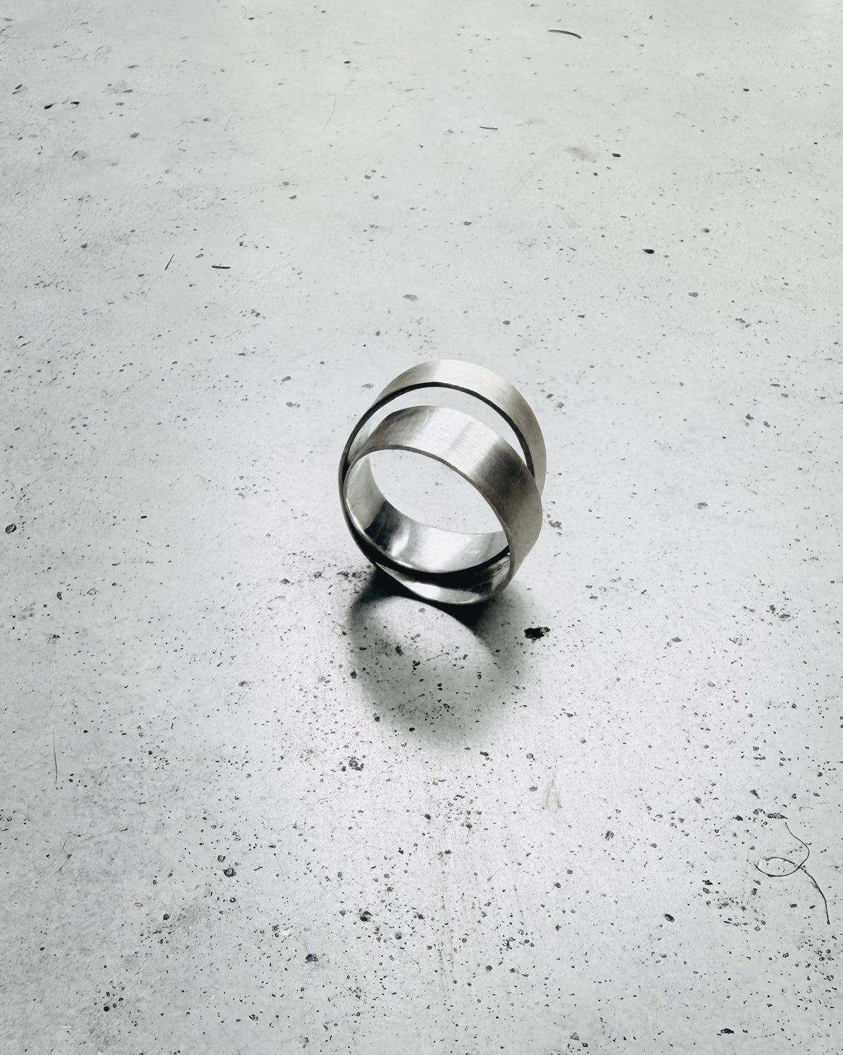 A polished silver-toned metal ring with a double-band design, standing upright on a textured gray concrete surface.