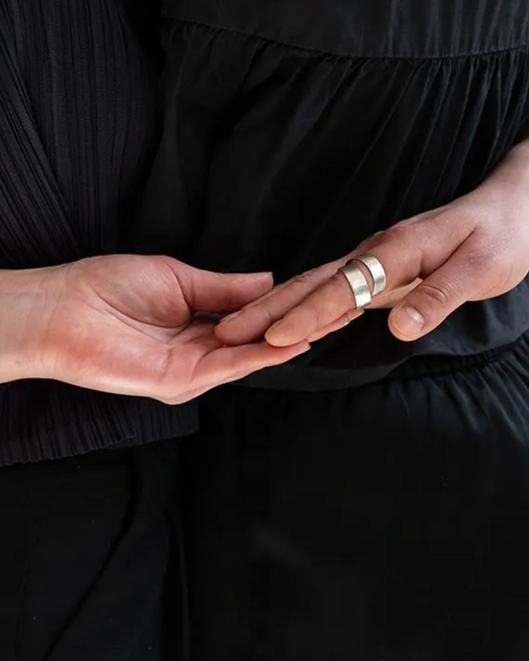 A close-up of a person wearing a wide, polished silver band ring on their finger, with their other hand resting gently over it. They are wearing a black pleated dress.