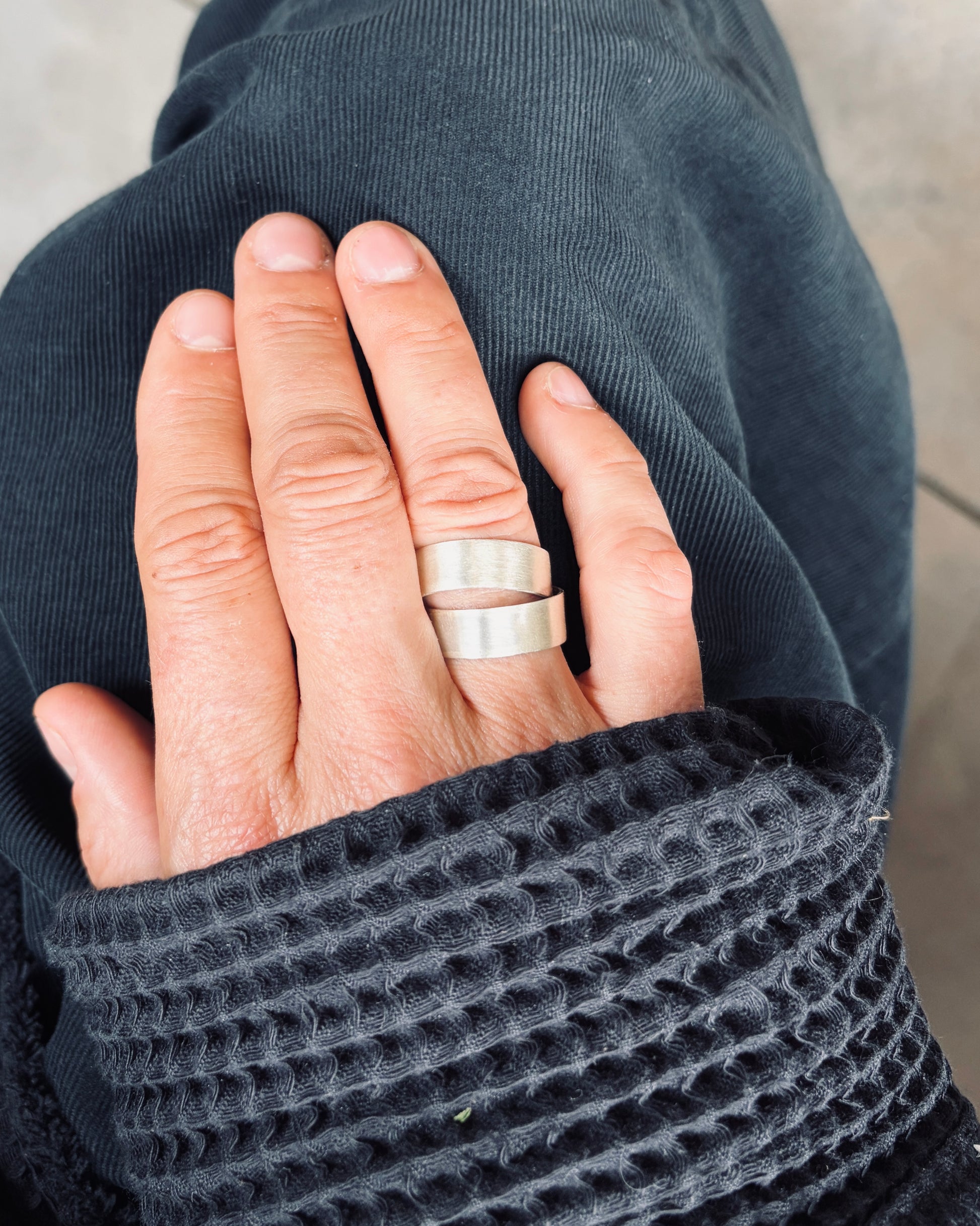 A close-up of a person's hand wearing two simple silver bands, resting on dark gray fabric.