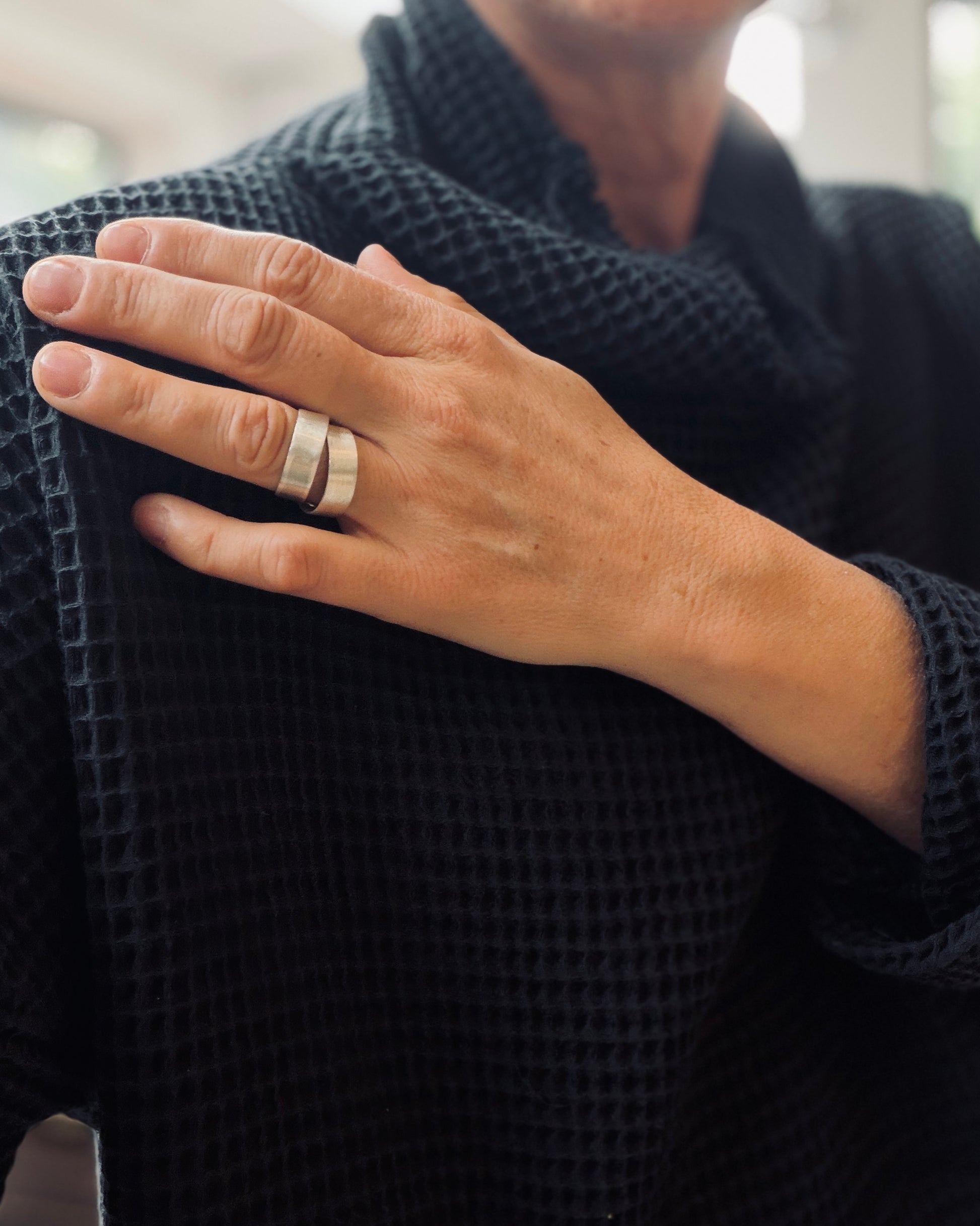 A close-up of a person's hand wearing two wide, polished silver bands, styled with a black waffle-knit sweater.