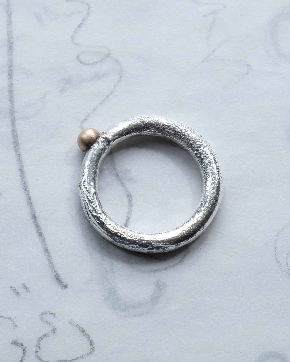 Close up of silver textured ring with brass ball sitting on top of tracing paper with faint pencil drawings.