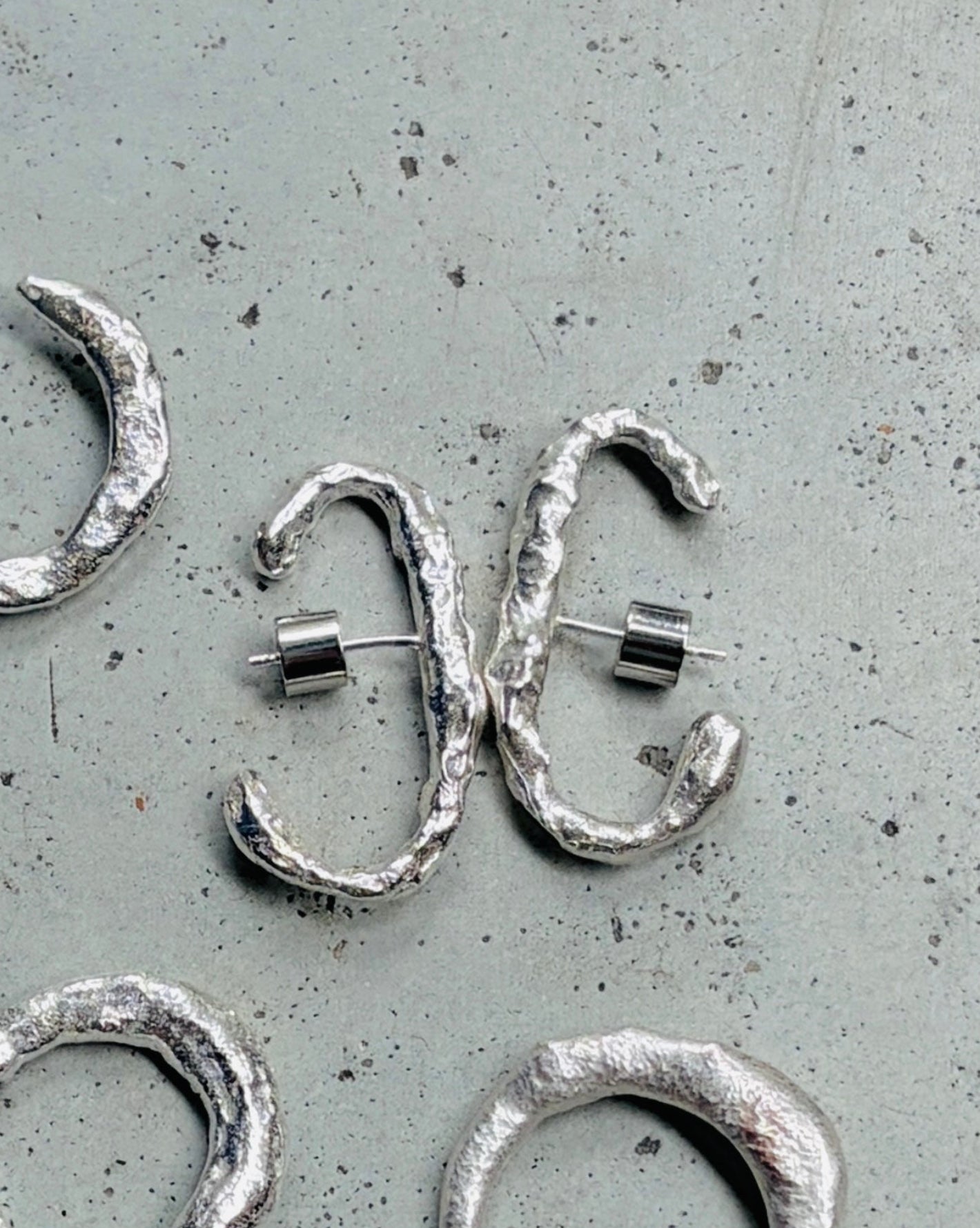 Five textured silver hoop earrings with asymmetrical wrap designs, displayed on a speckled gray surface.