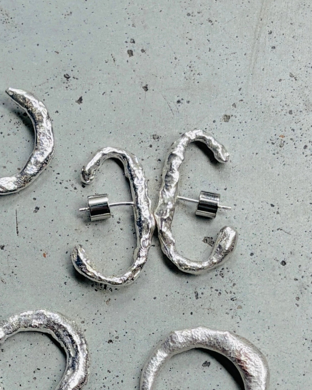 Five textured silver hoop earrings with asymmetrical wrap designs, displayed on a speckled gray surface.