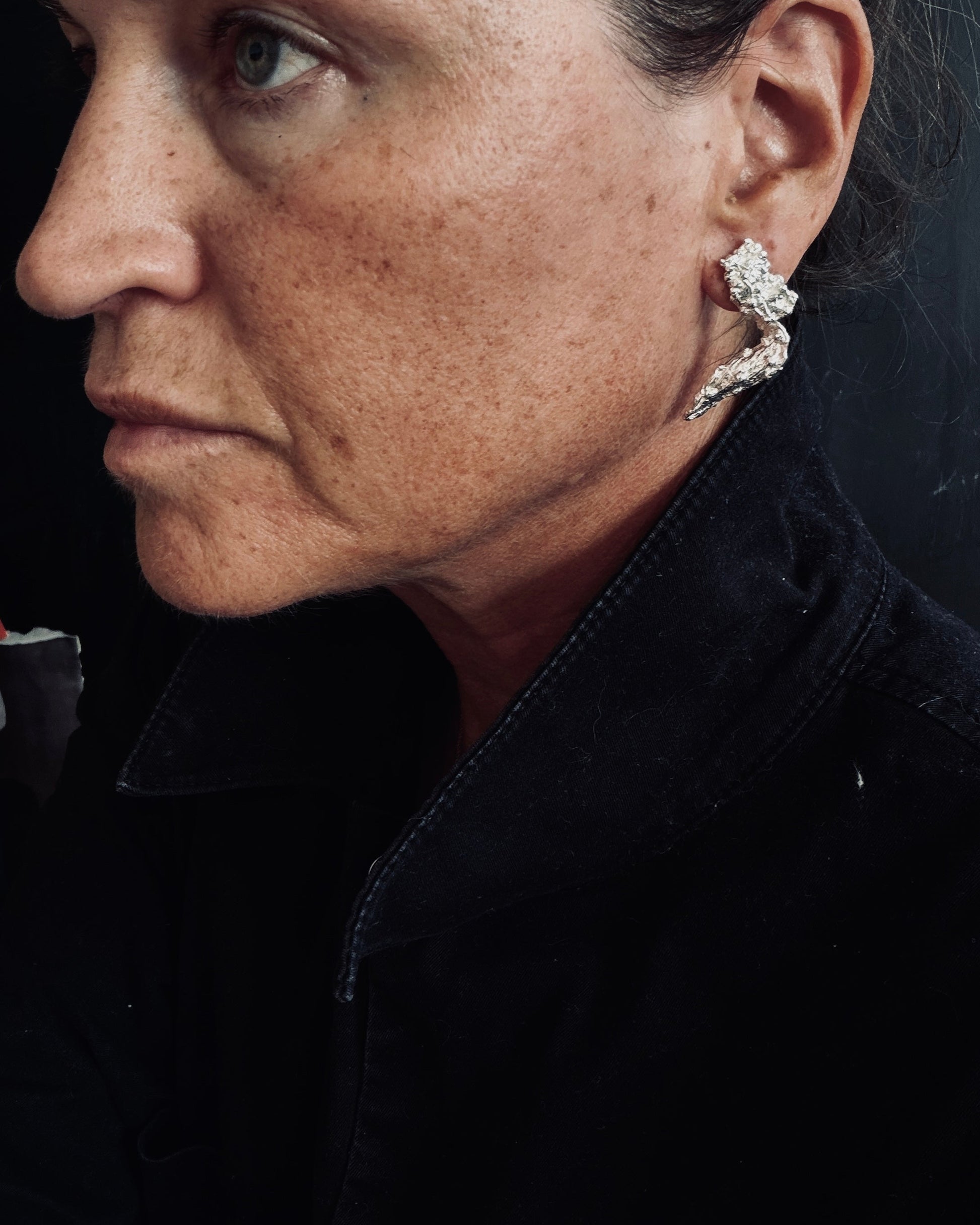 A close-up side profile of a woman wearing a sculptural, twisted silver-toned statement earring with clear gemstones, paired with a black jacket against a dark background.