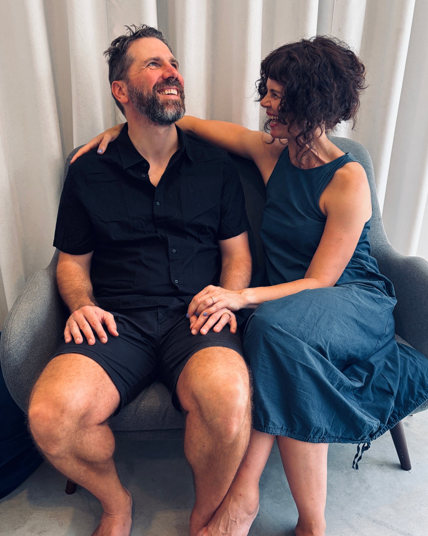 A man and a woman sitting together on a gray armchair in front of white curtains. The man wears a black short-sleeve button-up shirt and black shorts, while the woman wears a sleeveless dark teal dress.