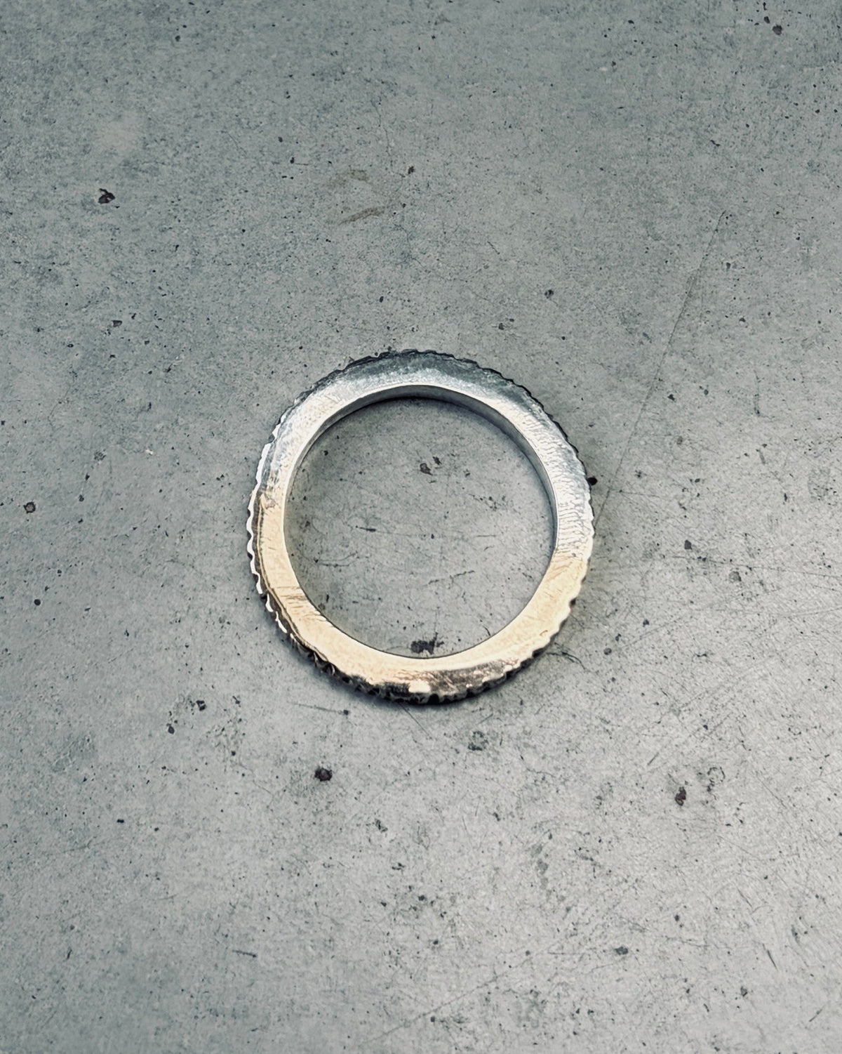 A handmade silver-toned metal ring with a textured, rough-washed finish and a small notch on one side, placed on a gray concrete surface.