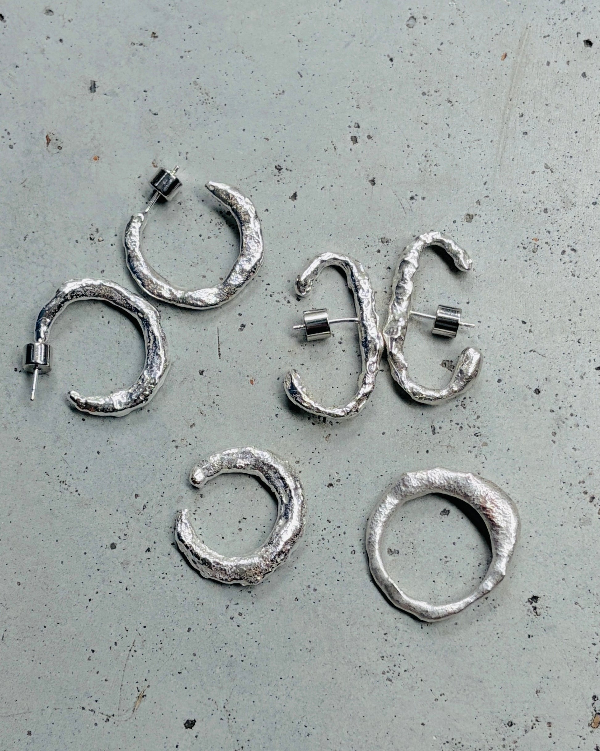 Five textured silver hoop earrings with asymmetrical wrap designs, displayed on a speckled gray surface.