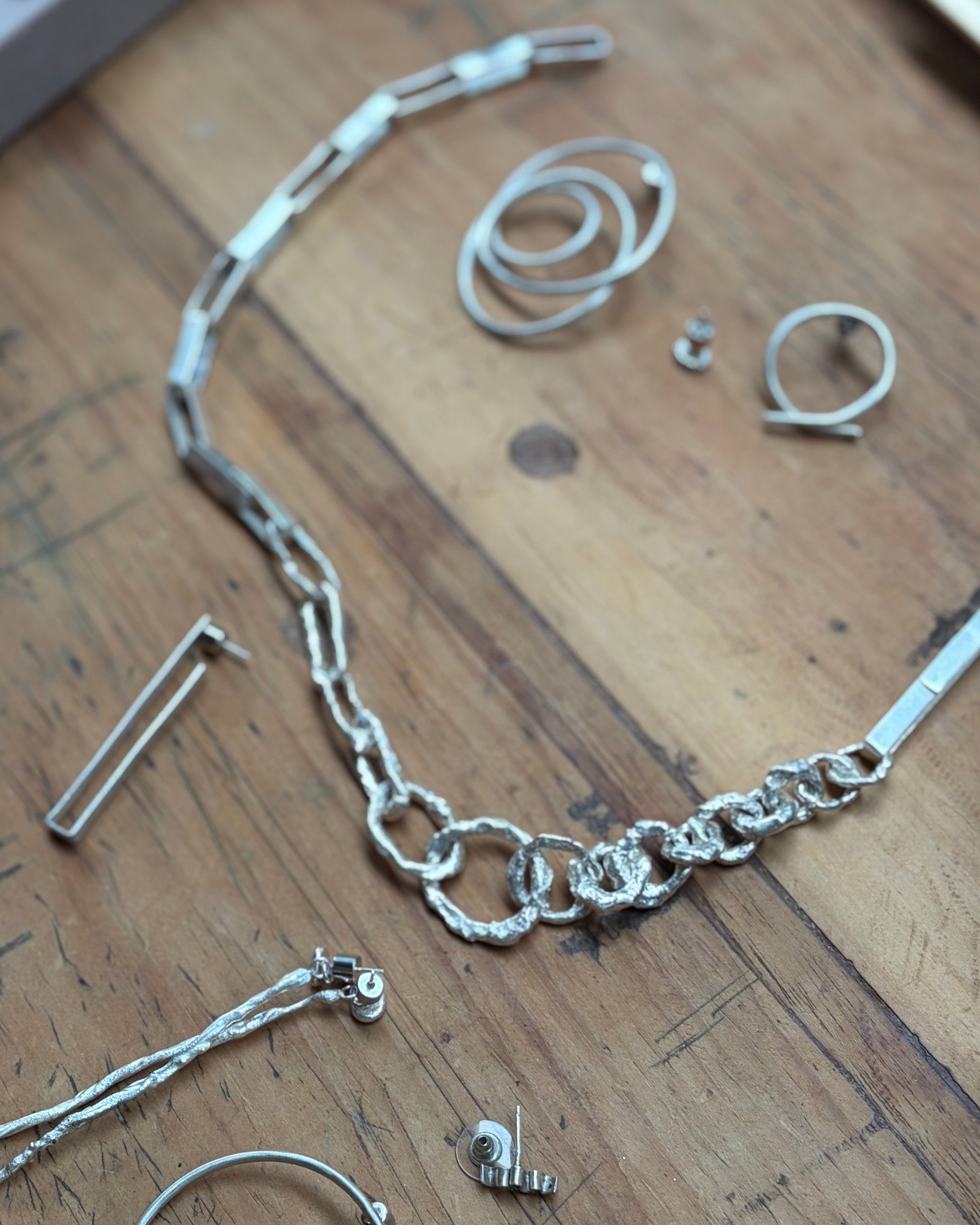 A set of silver-toned jewelry components, including a chain bracelet, rings, a pair of dangling earrings, and a hook clasp, laid out on a rustic wooden surface.