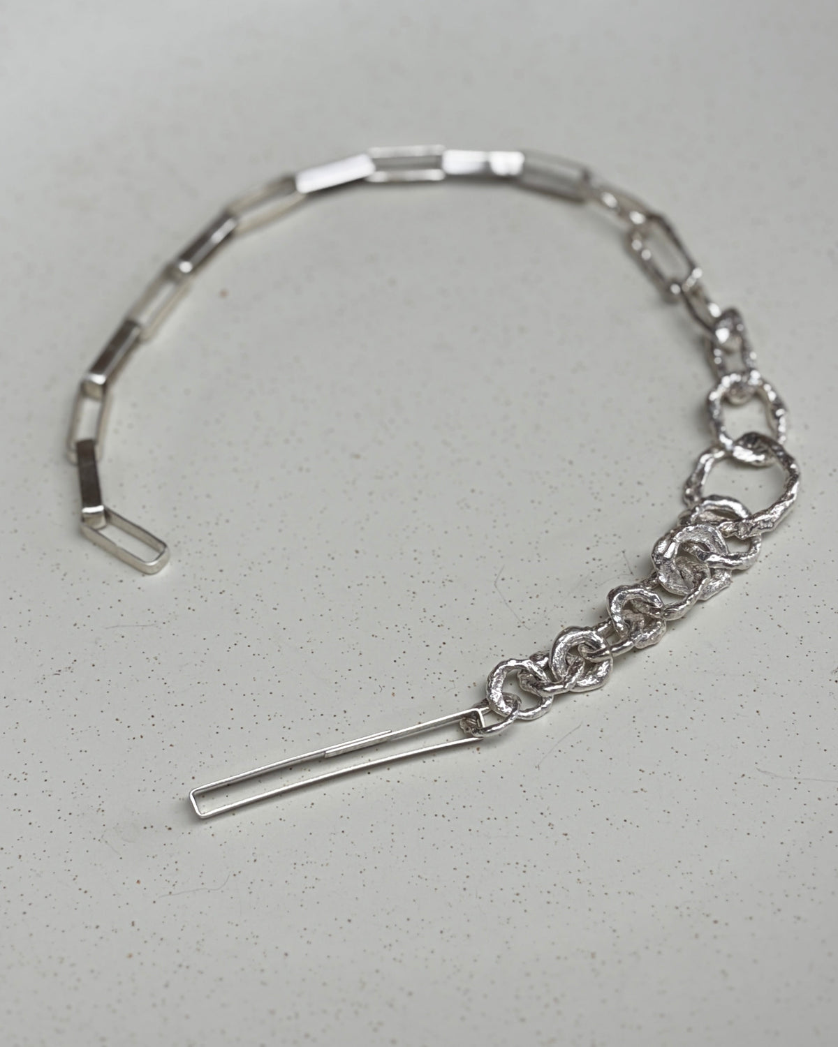 A silver-toned metal bracelet with a combination of a flat segmented band and a delicate chain link section, resting on a white ceramic plate.