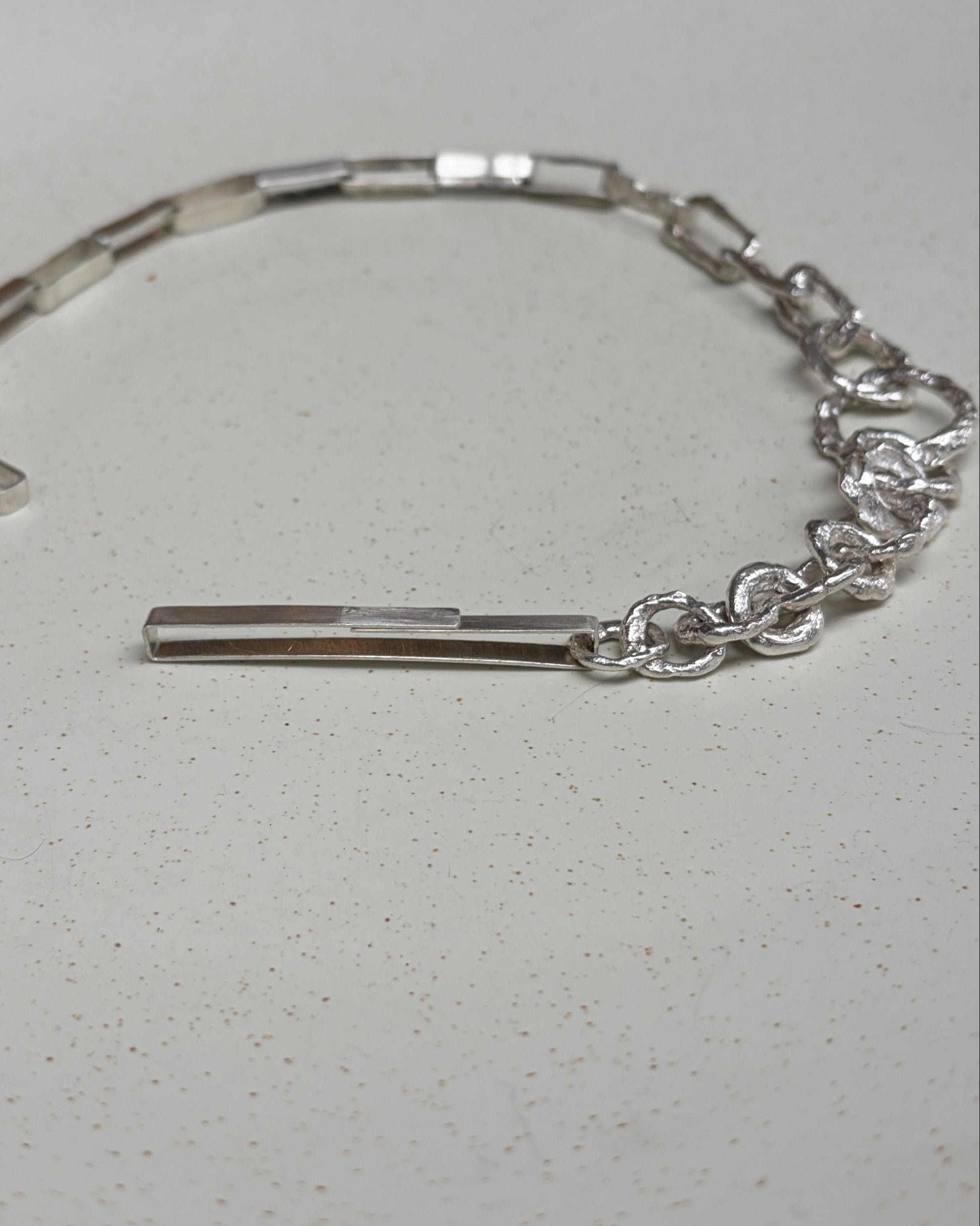 A silver-toned metal bracelet with a link chain and a vertical bar clasp, displayed on a white speckled surface.
