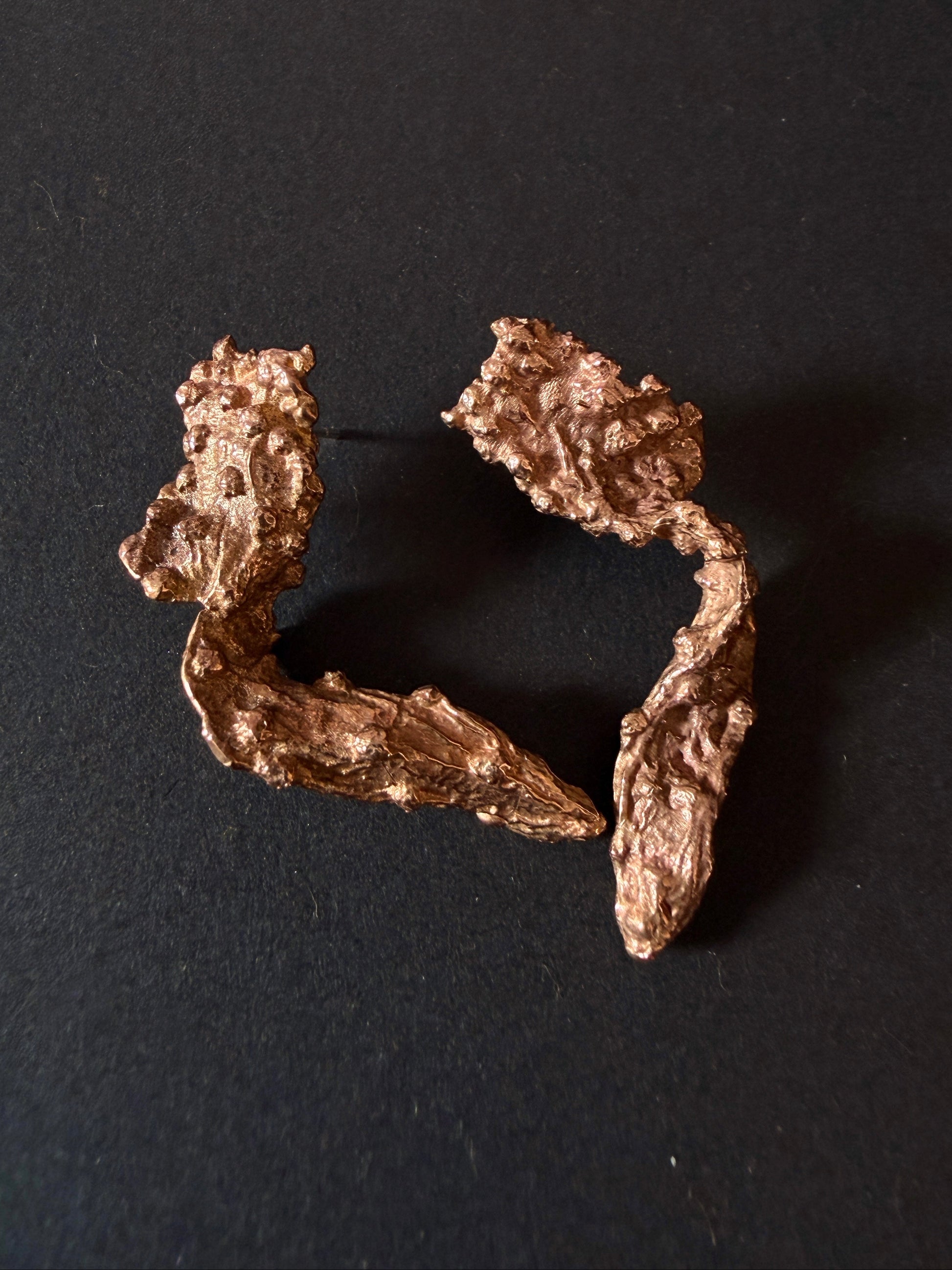 A pair of organic-shaped metallic earrings with a textured, hammered finish in a warm copper or bronze tone, displayed on a dark black background.