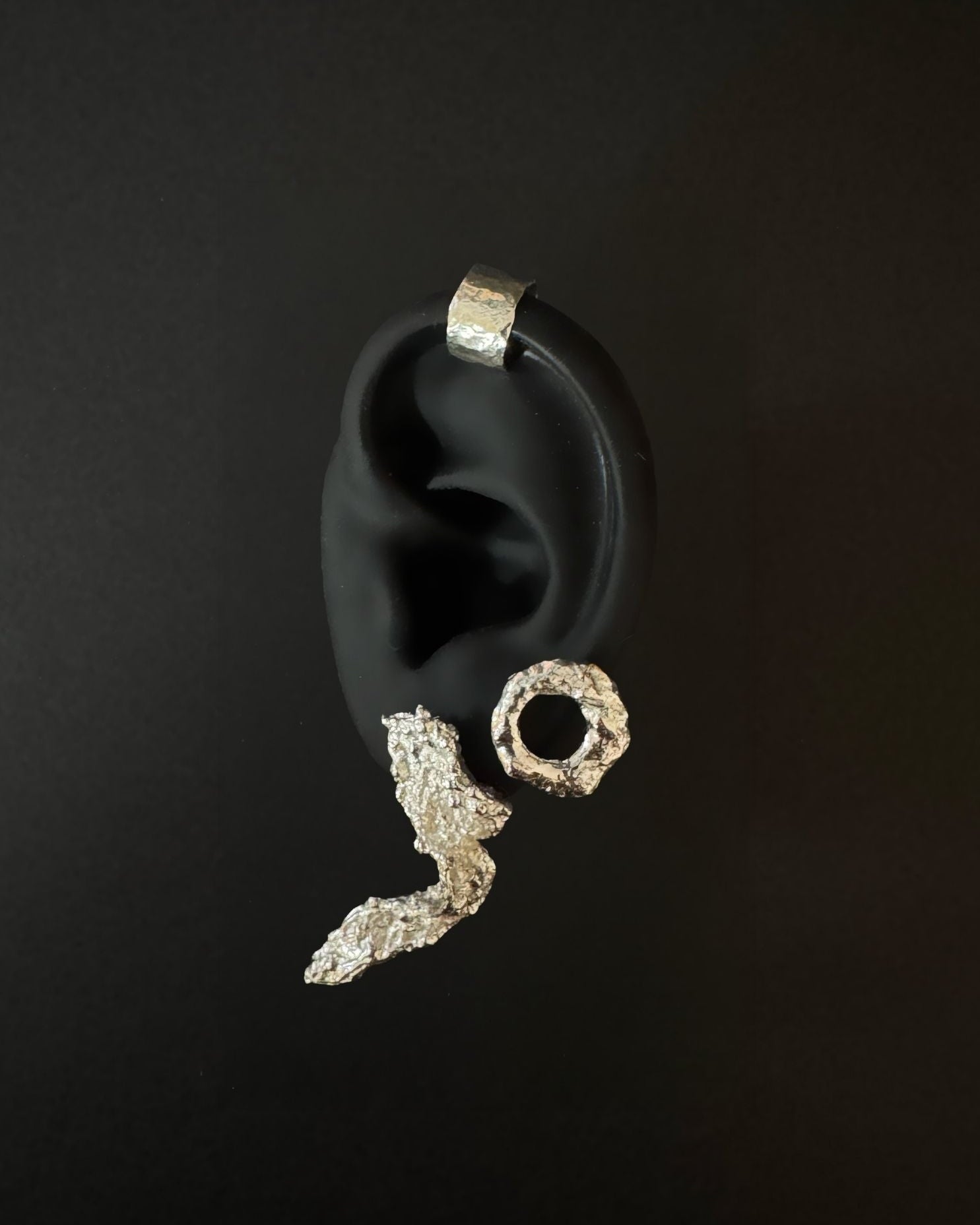 A pair of textured silver earrings, including a lightning bolt stud and a circular hoop, displayed on a black ear form against a dark background.