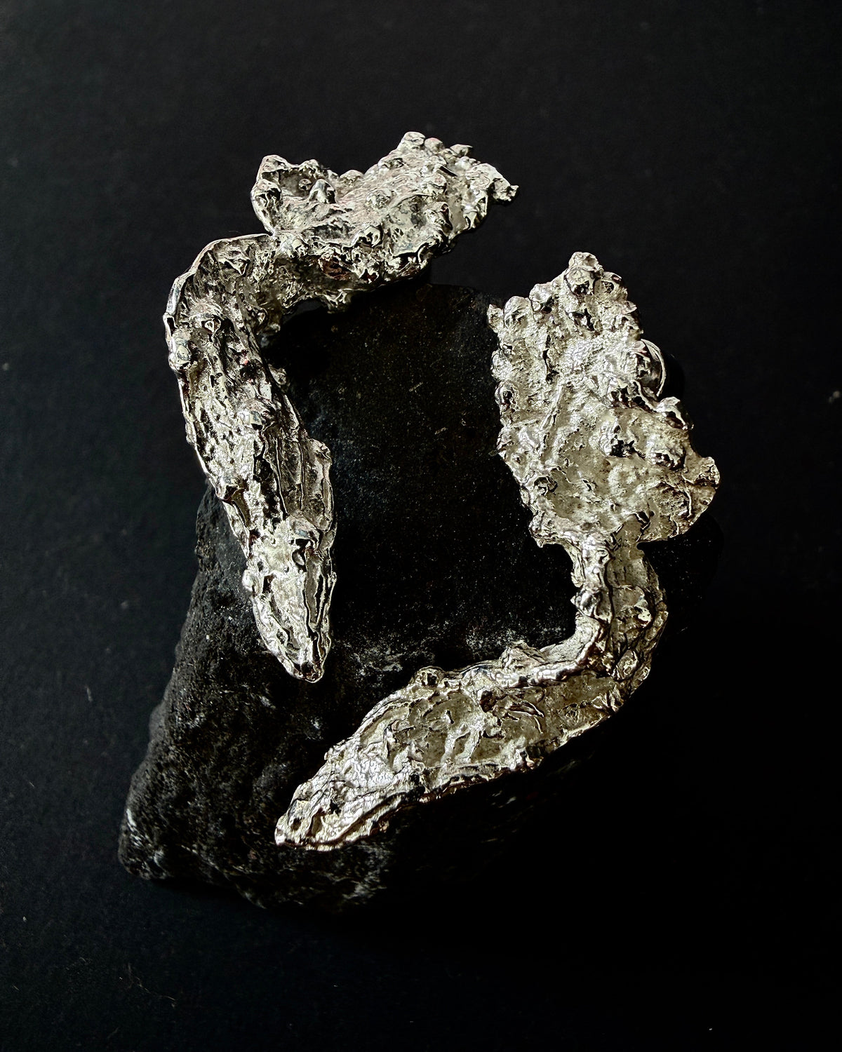 A raw, rough-textured silver metallic specimen, resembling a mineral vein or natural ore, displayed on a dark black background.