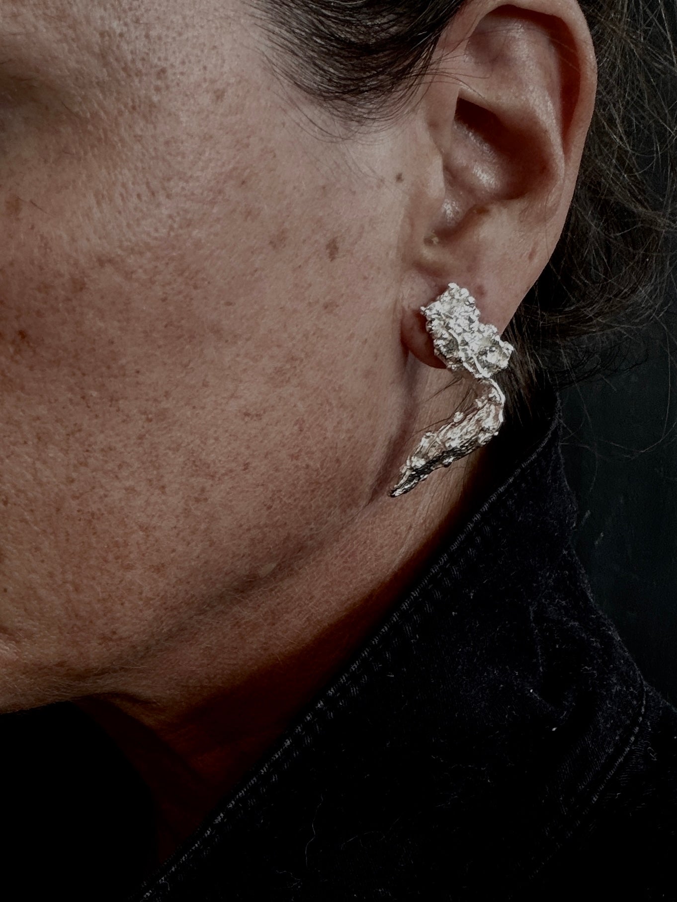 A close-up of a person's ear wearing a textured, organic-shaped silver earring against a dark background.
