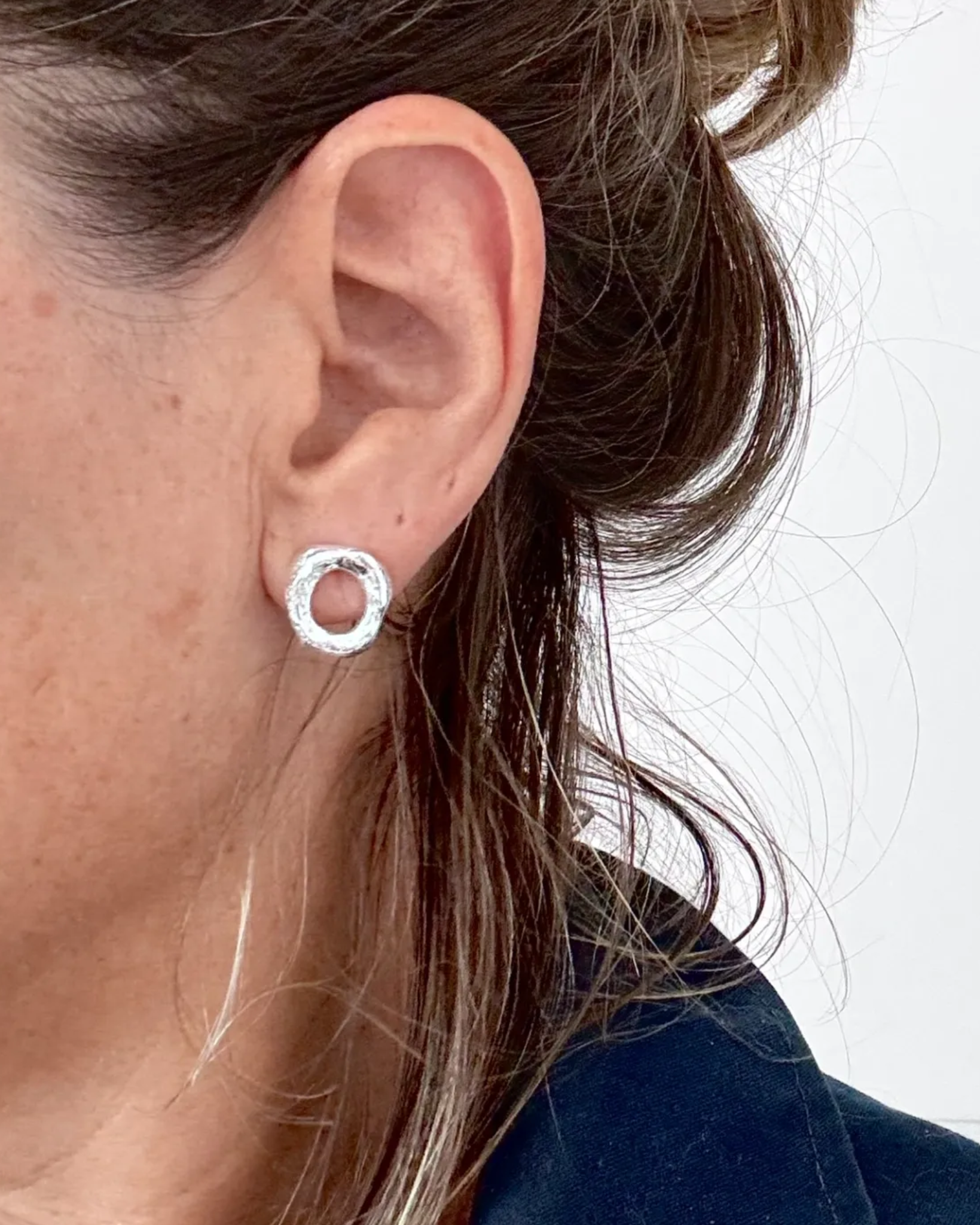 A close-up of a person's ear wearing a small silver hoop earring with a textured, hammered finish.