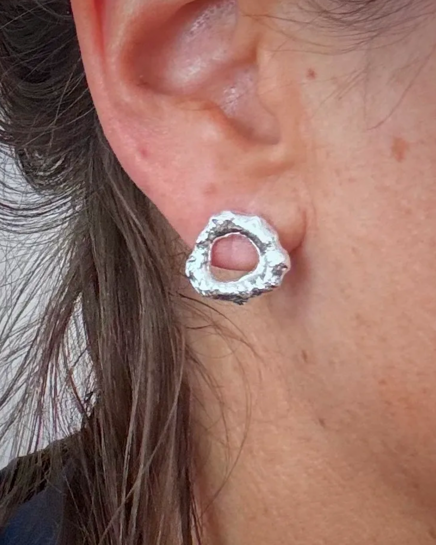 A close-up of a person's ear wearing a textured, irregularly shaped silver hoop earring.