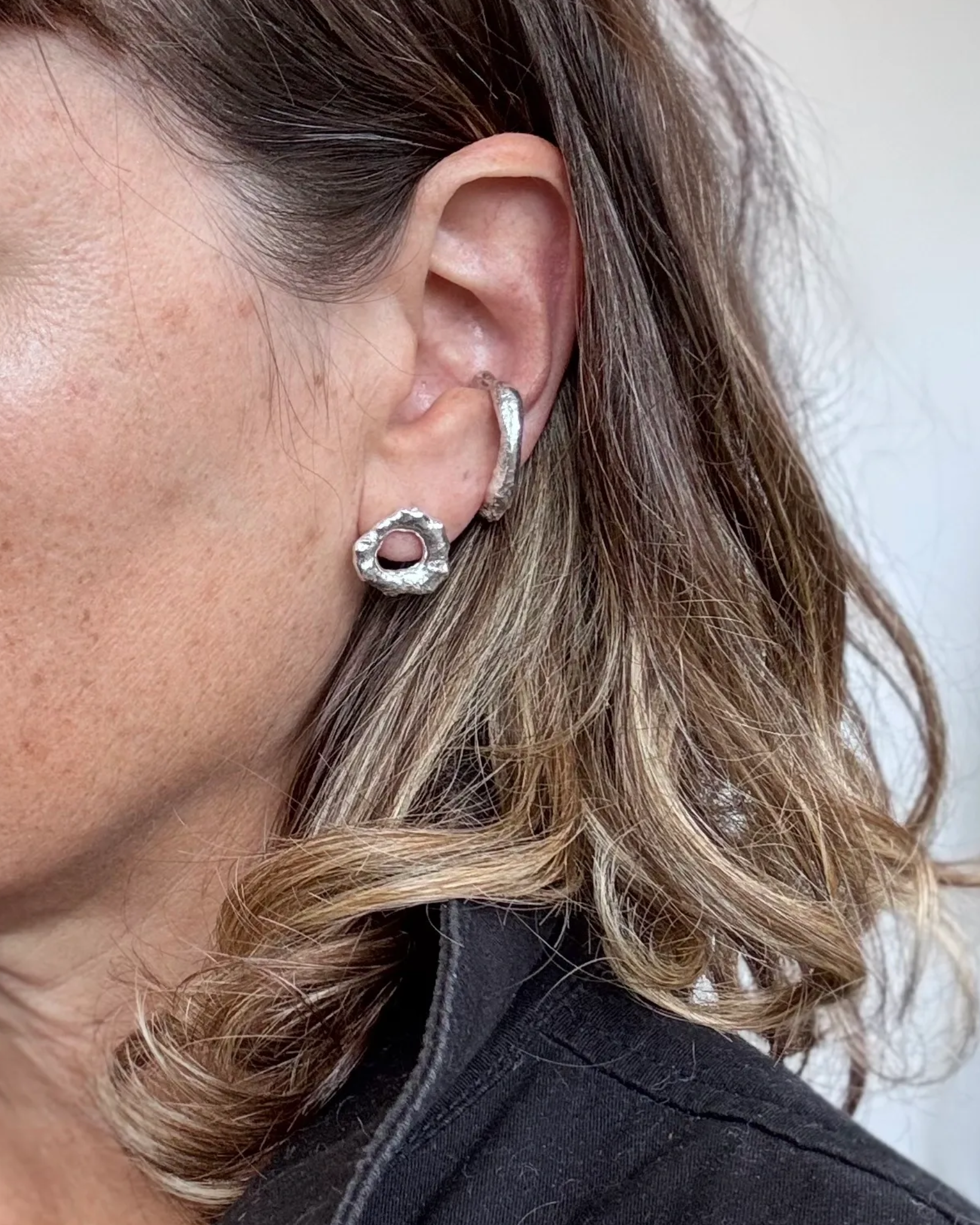 A close-up of a woman's ear wearing a silver-toned double-hoop earring and a textured silver ring stud with a pink gemstone.