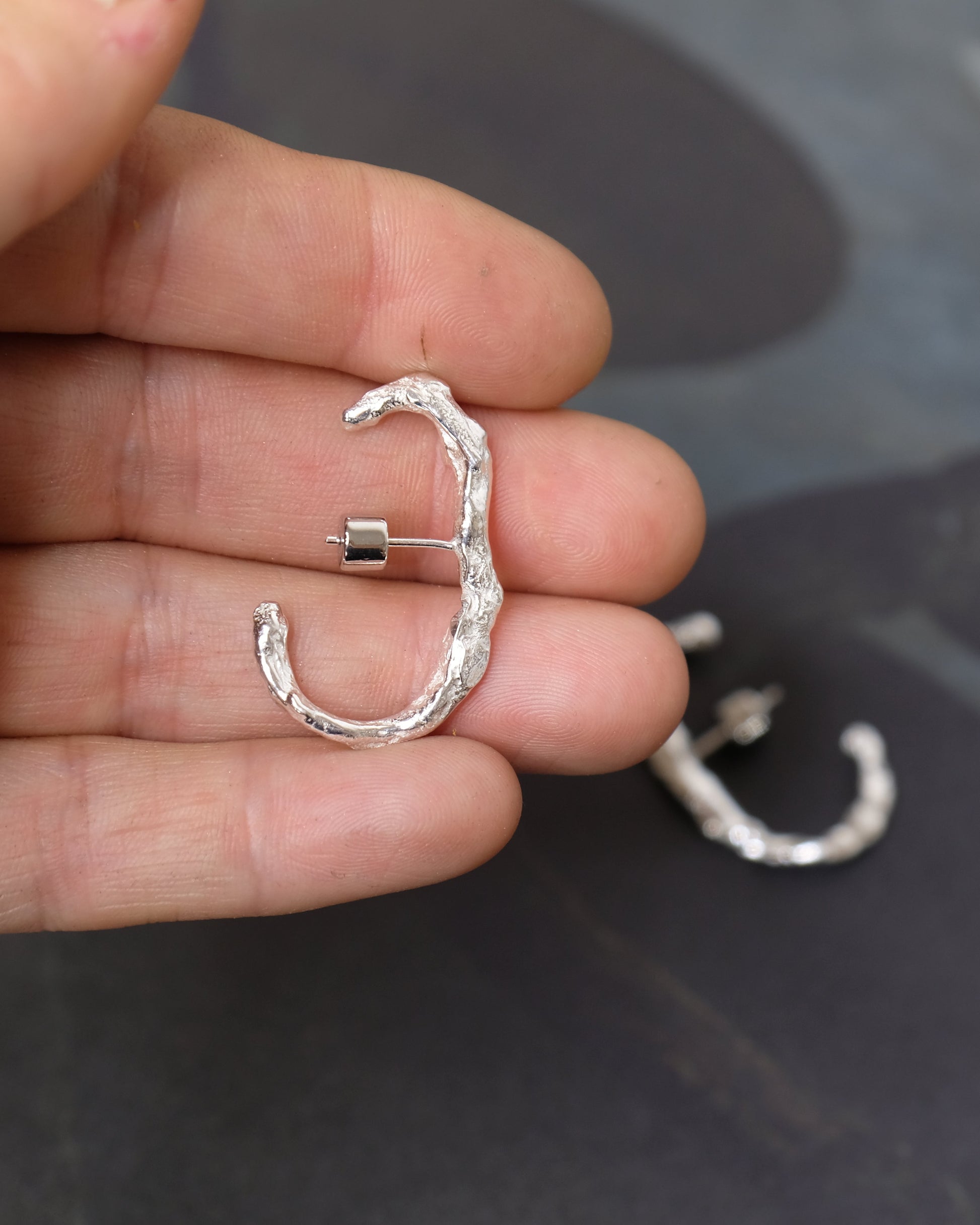A hand holding a small, textured silver C-shaped earring, with a second matching earring resting on a dark surface in the background.