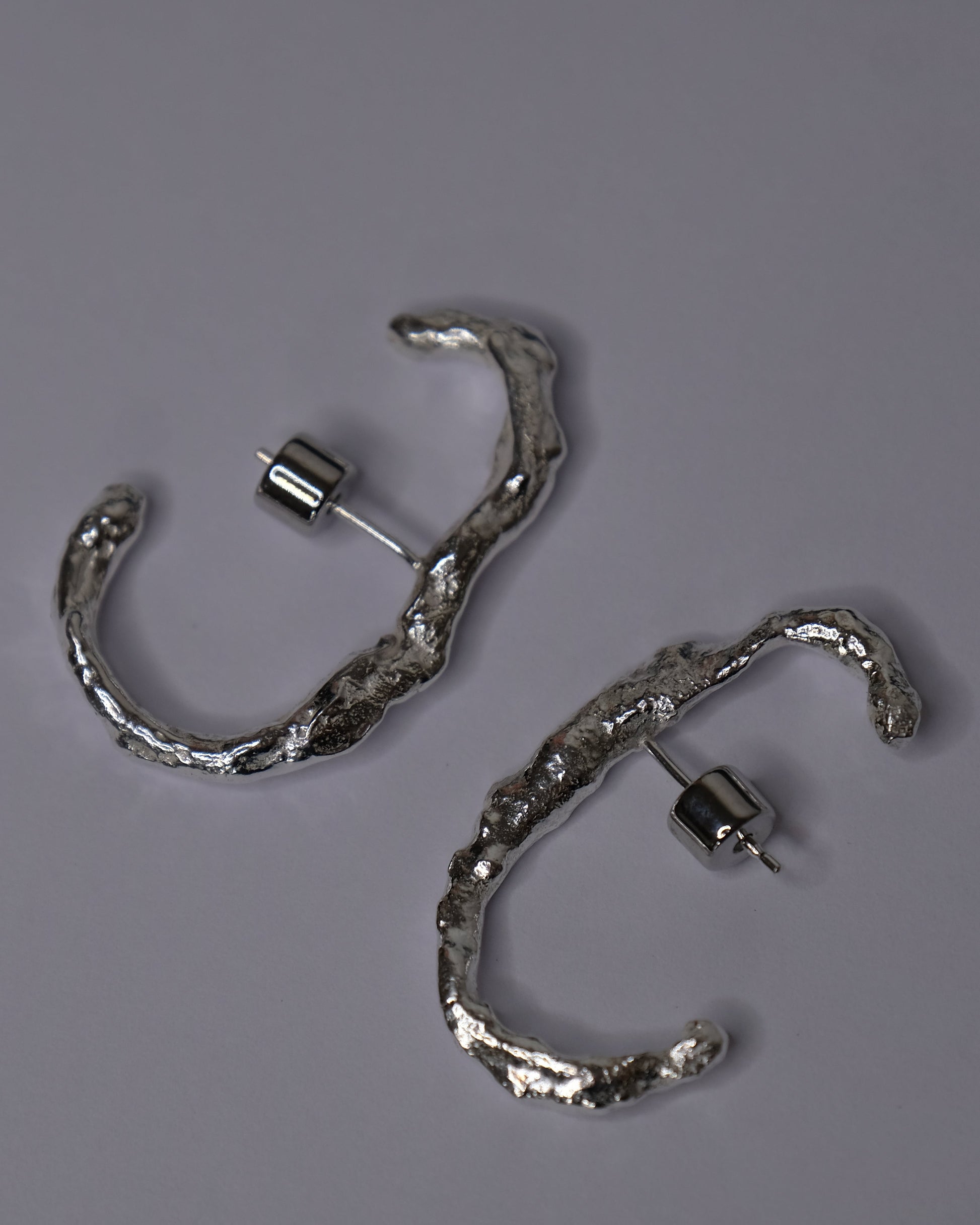 A pair of textured silver hoop earrings with a hammered, organic finish, displayed on a neutral gray background.
