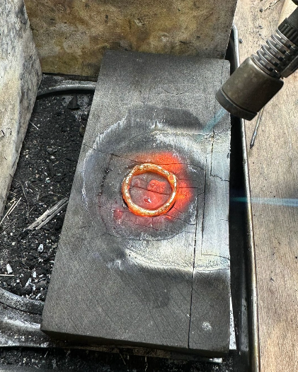 Charcoal block with molten ring being melted with torch pointing towards block.