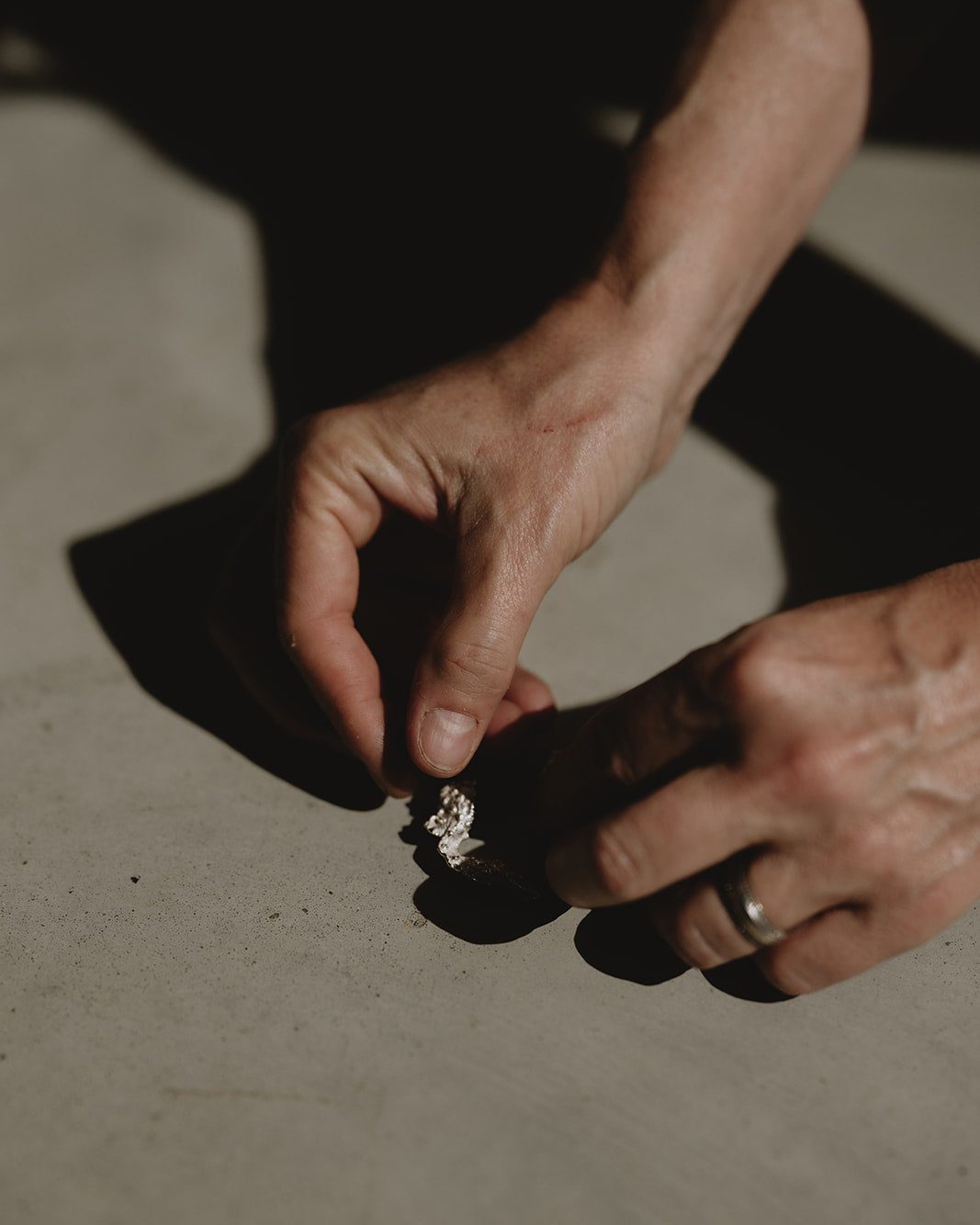 Close-up of a person's hands holding a small silver ring on a textured concrete surface under dramatic, high-contrast lighting.