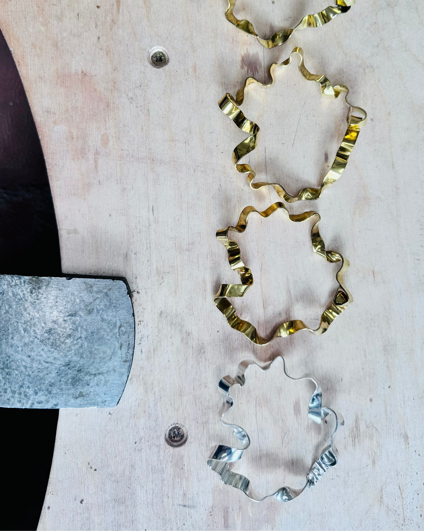 Four ruffled metal cookie cutters in gold, bronze, and silver finishes arranged vertically on a light-colored wooden board, with a metal mallet handle visible on the left.