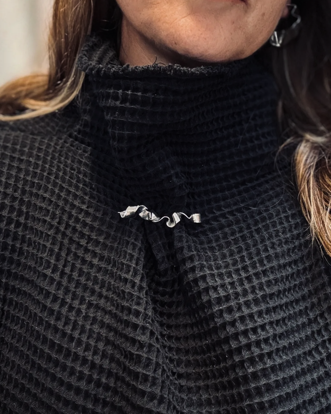 A close-up of a person wearing a black waffle-knit turtleneck sweater with a silver metallic wavy brooch at the center of the chest.