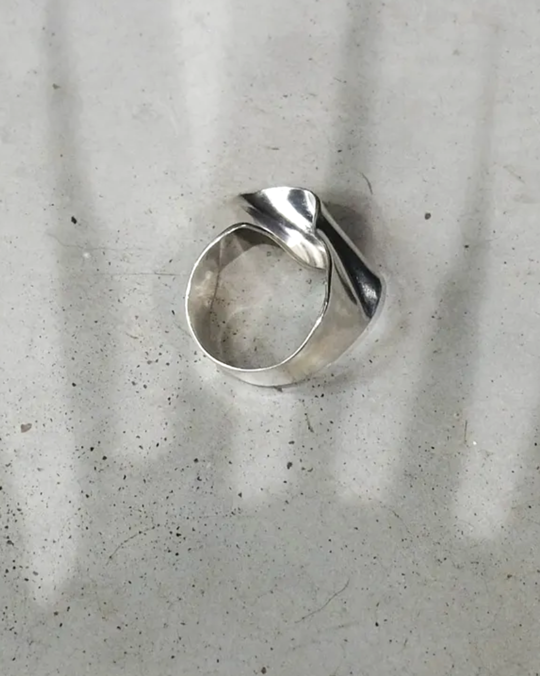 A polished silver ring with a twisted, organic shape, resting on a textured light gray surface with soft shadows.