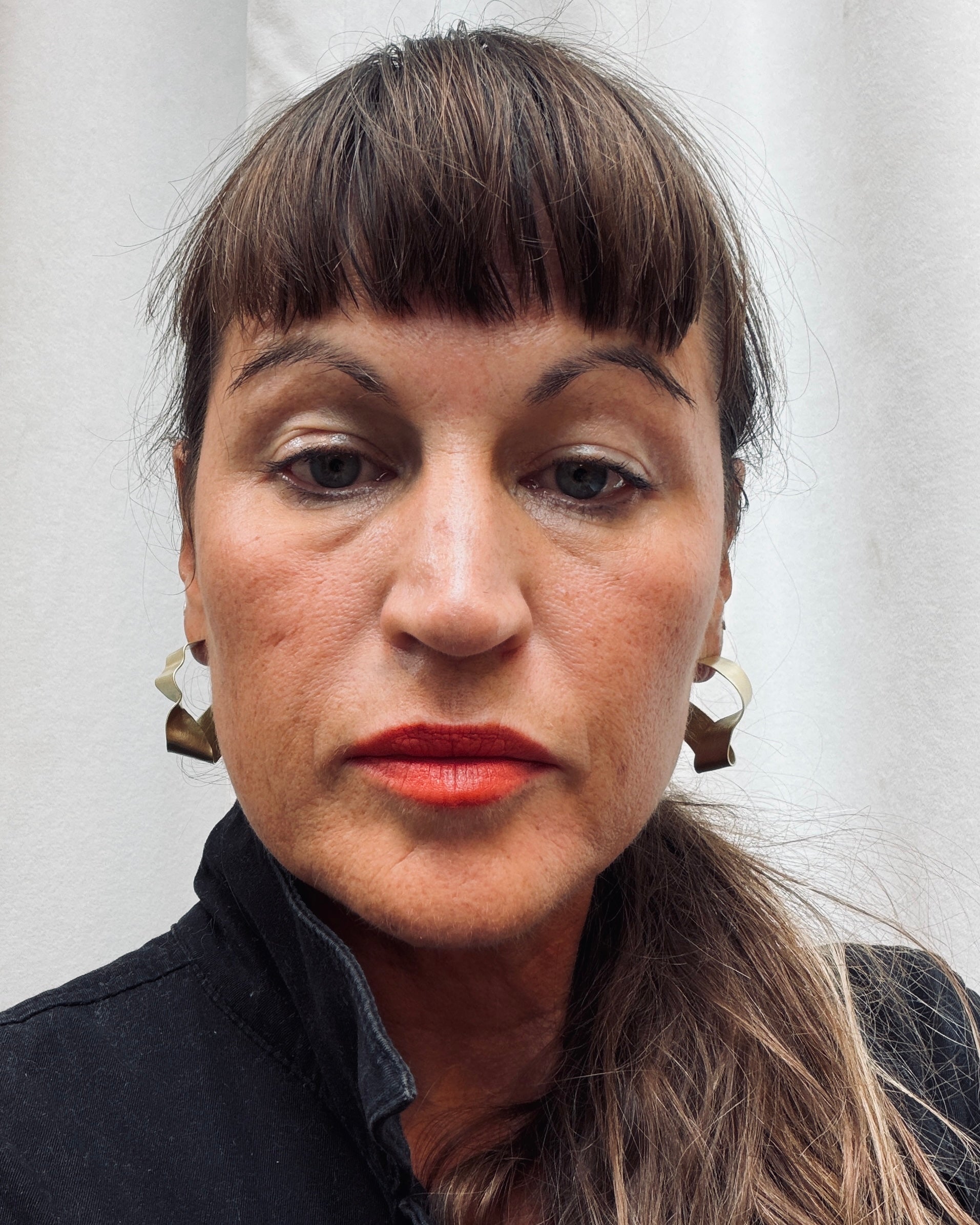A close-up portrait of a woman with dark brown hair, bangs, and red lipstick, wearing gold geometric earrings and a dark jacket against a light gray background.