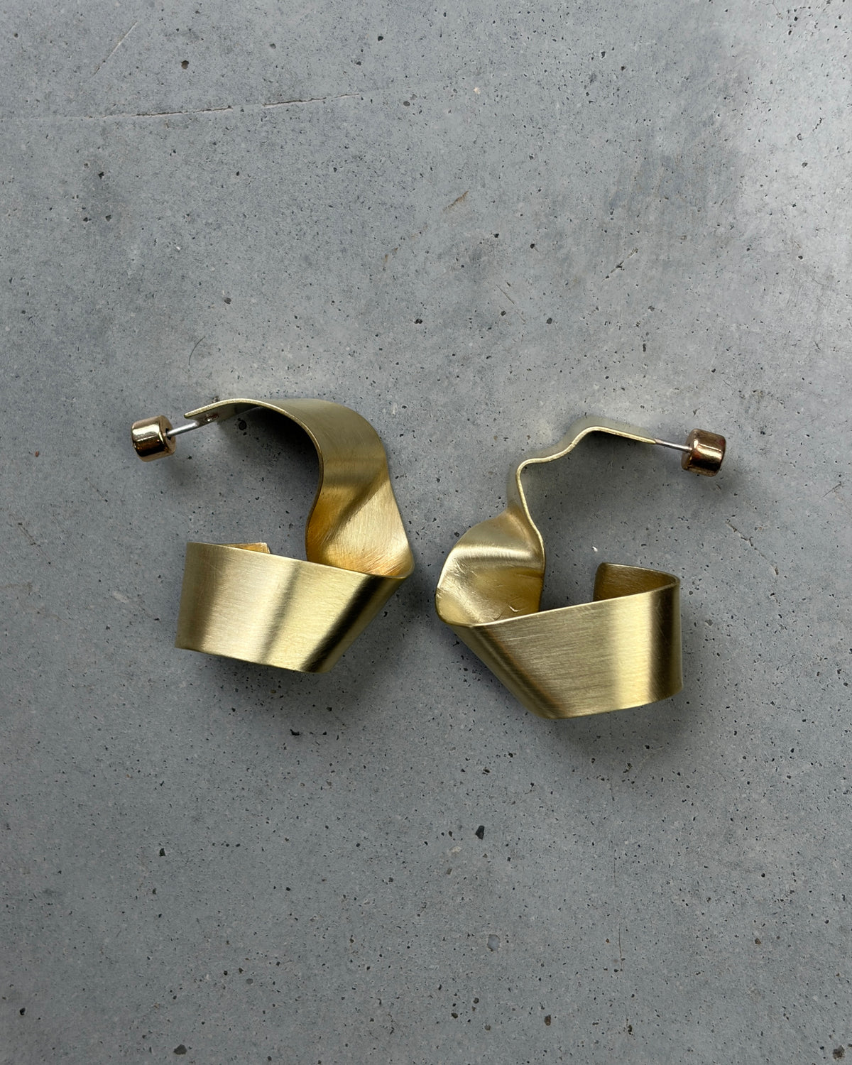 A pair of abstract, open hoop earrings made of brushed brass, displayed on a textured gray concrete surface.