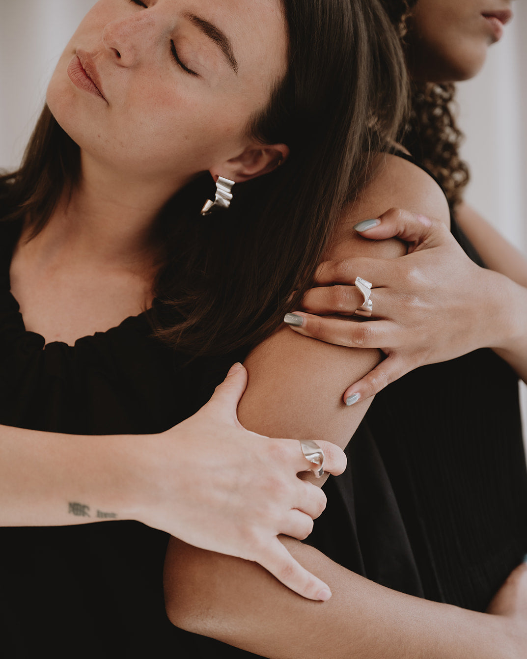 Close-up of a woman in a black top receiving a shoulder massage, with another person's hands visible on her upper arm. She is wearing silver hoop earrings and rings.