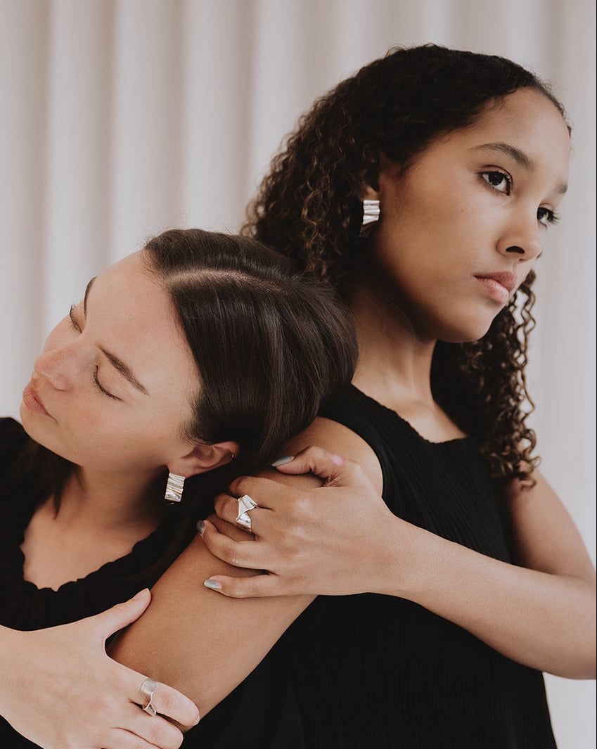 Two women embracing in a black dress, wearing silver geometric stud earrings and rings, set against a soft beige curtain background.