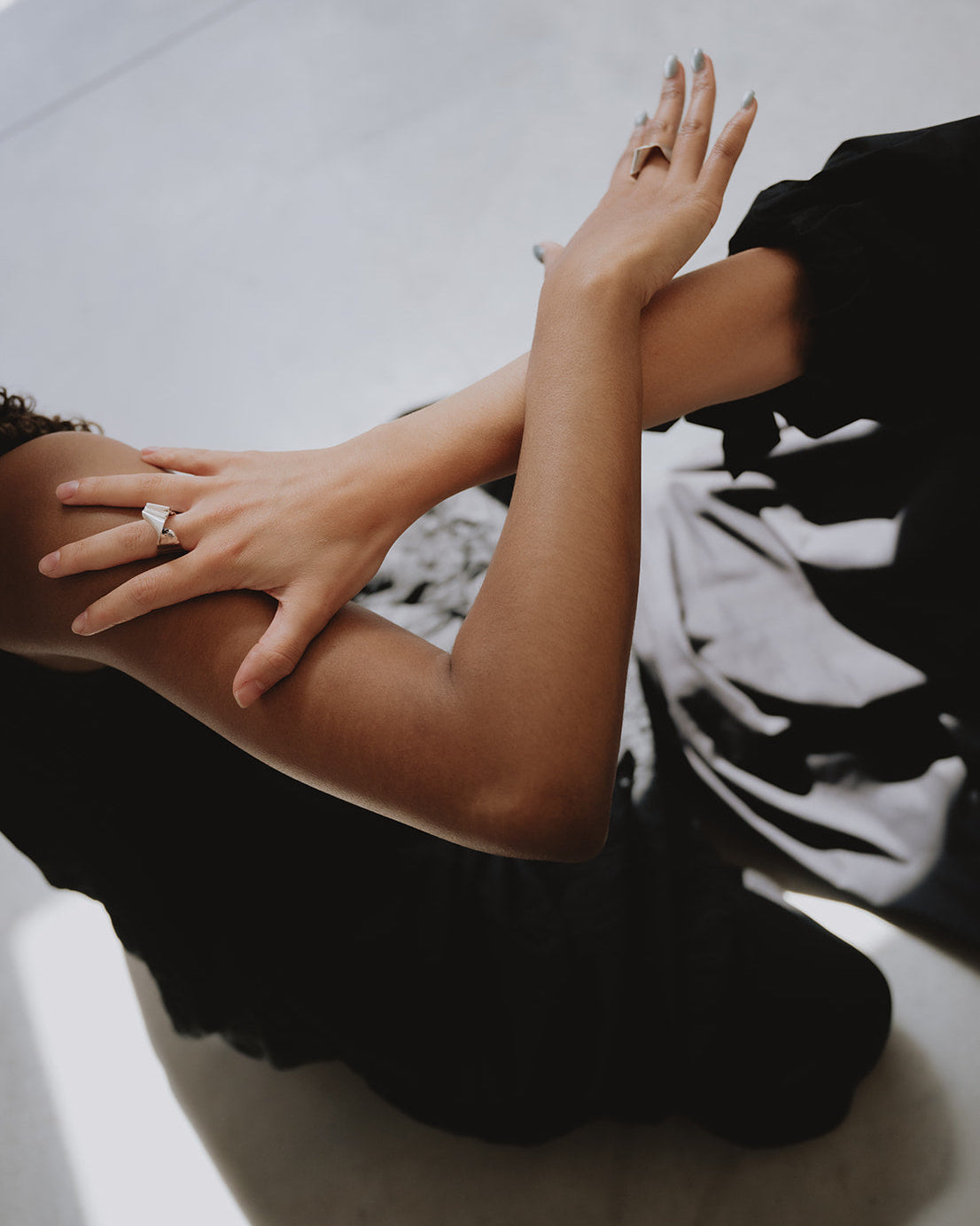 A close-up lifestyle shot of a person wearing a black dress, showing their arm and hand resting on another person's shoulder. The person in the black dress is wearing a silver ring with a clear gemstone on their finger.
