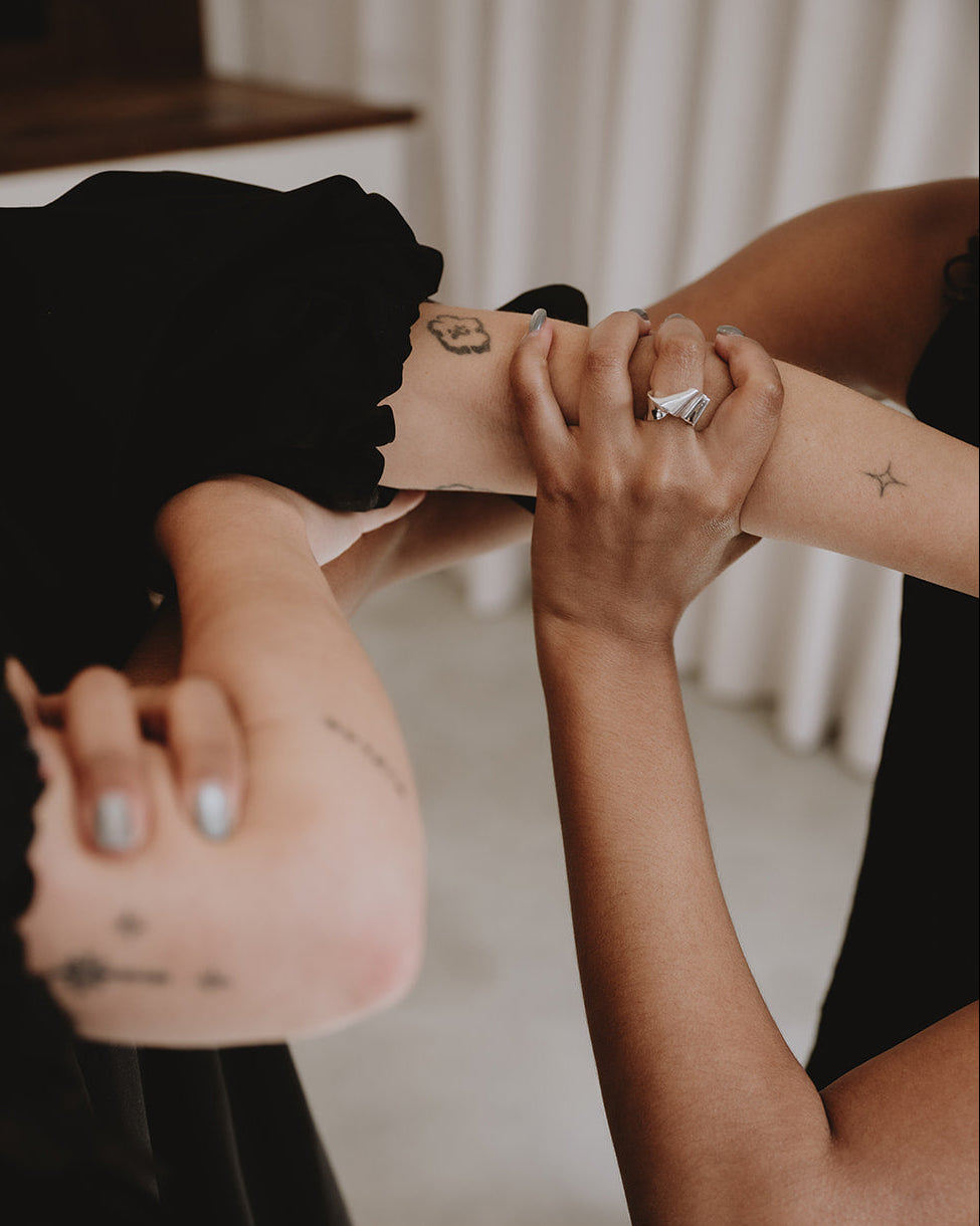 A close-up lifestyle shot of a person applying a small black tattoo or temporary tattoo design onto another person's forearm. The scene is shot in soft, warm indoor lighting with a blurred background of neutral curtains and a light-colored floor.