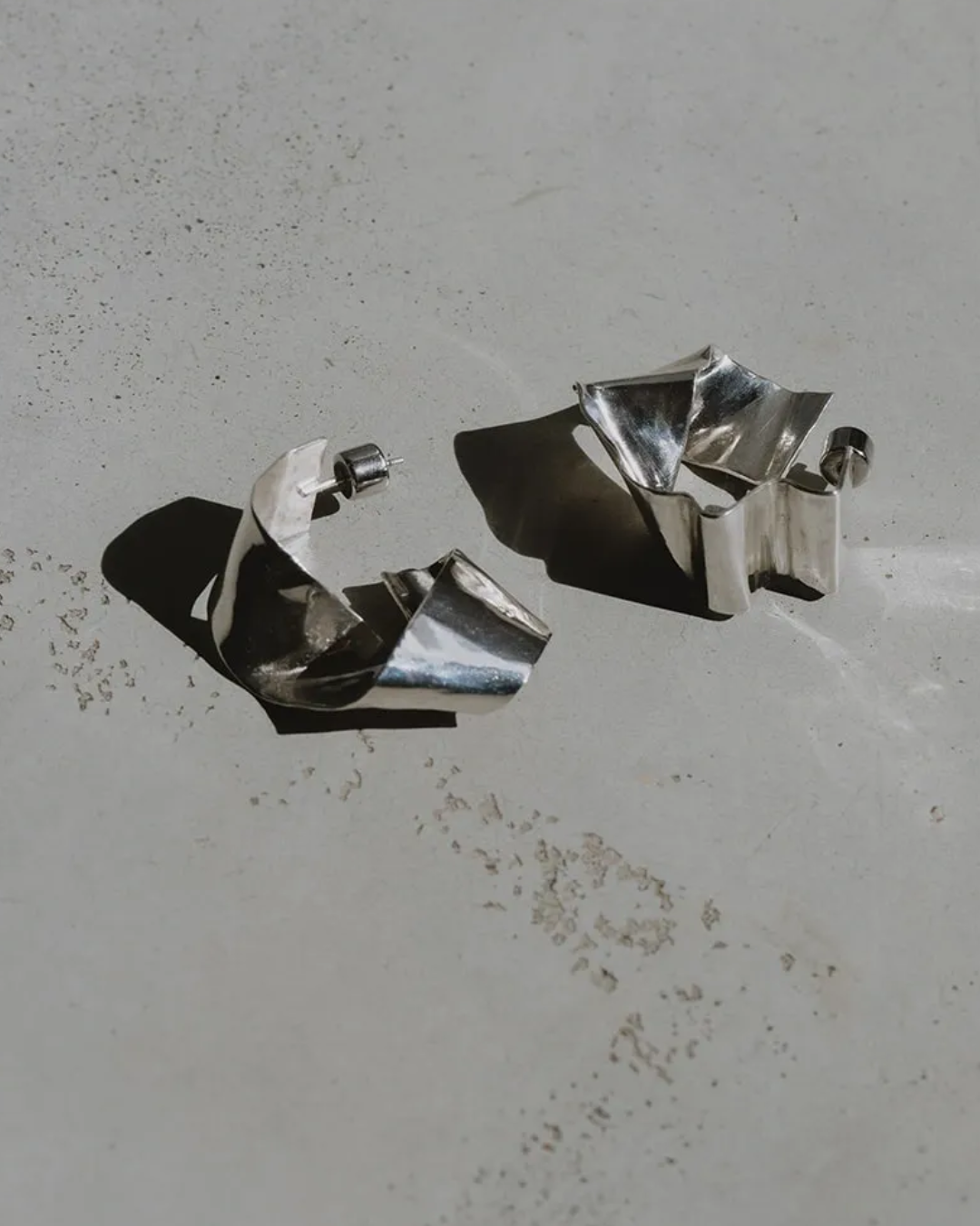A pair of sculptural, crumpled metallic silver earrings displayed on a textured light gray surface with sharp shadows.