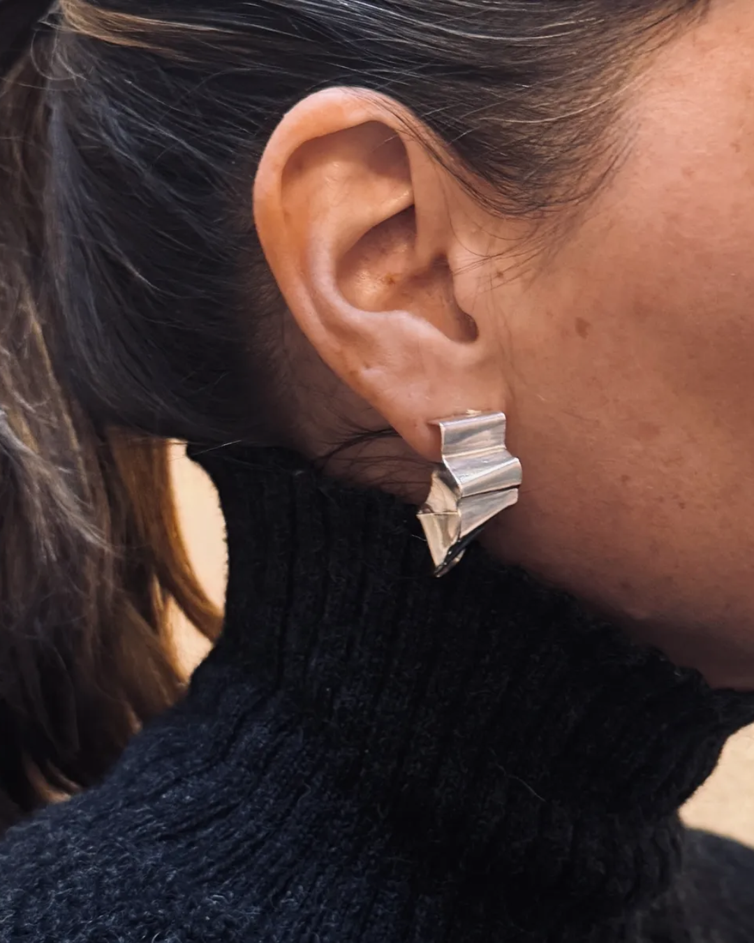 A close-up of a person's ear wearing a chunky, geometric silver earring with layered, stepped triangular facets.