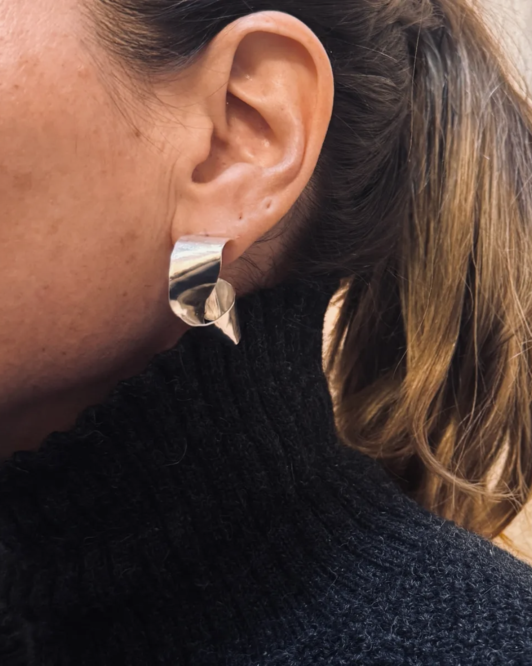 A close-up of a person's ear wearing a chunky, polished silver hoop earring with a textured, layered design.