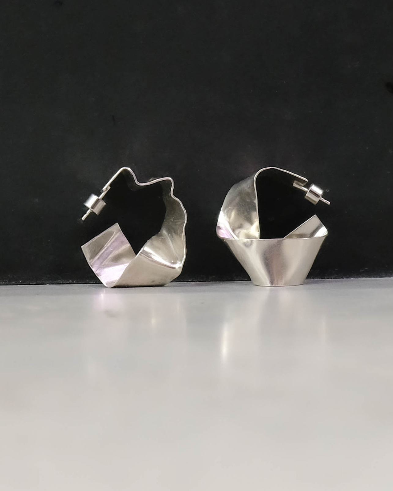 pair of sculptural silver hoops sitting against black wall on concrete floor