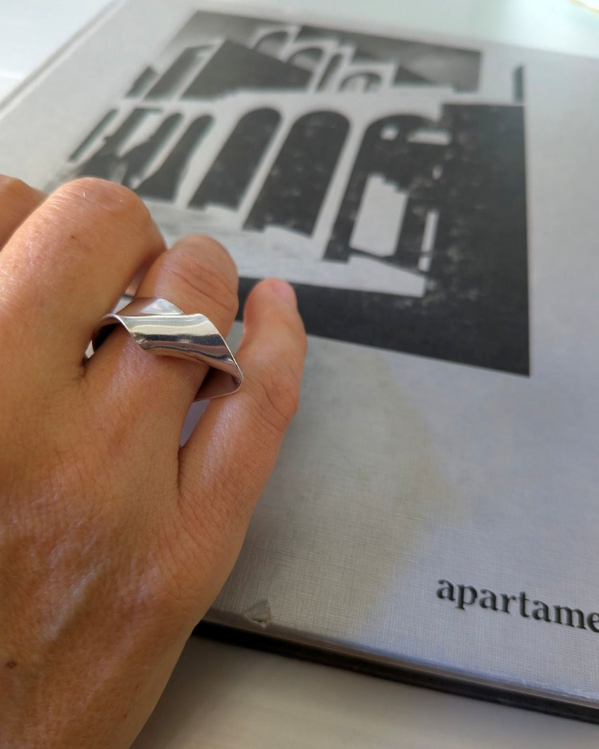 A close-up of a hand wearing a wide, polished silver ring with a geometric, stepped triangular design, resting on a gray book with black and white architectural imagery.