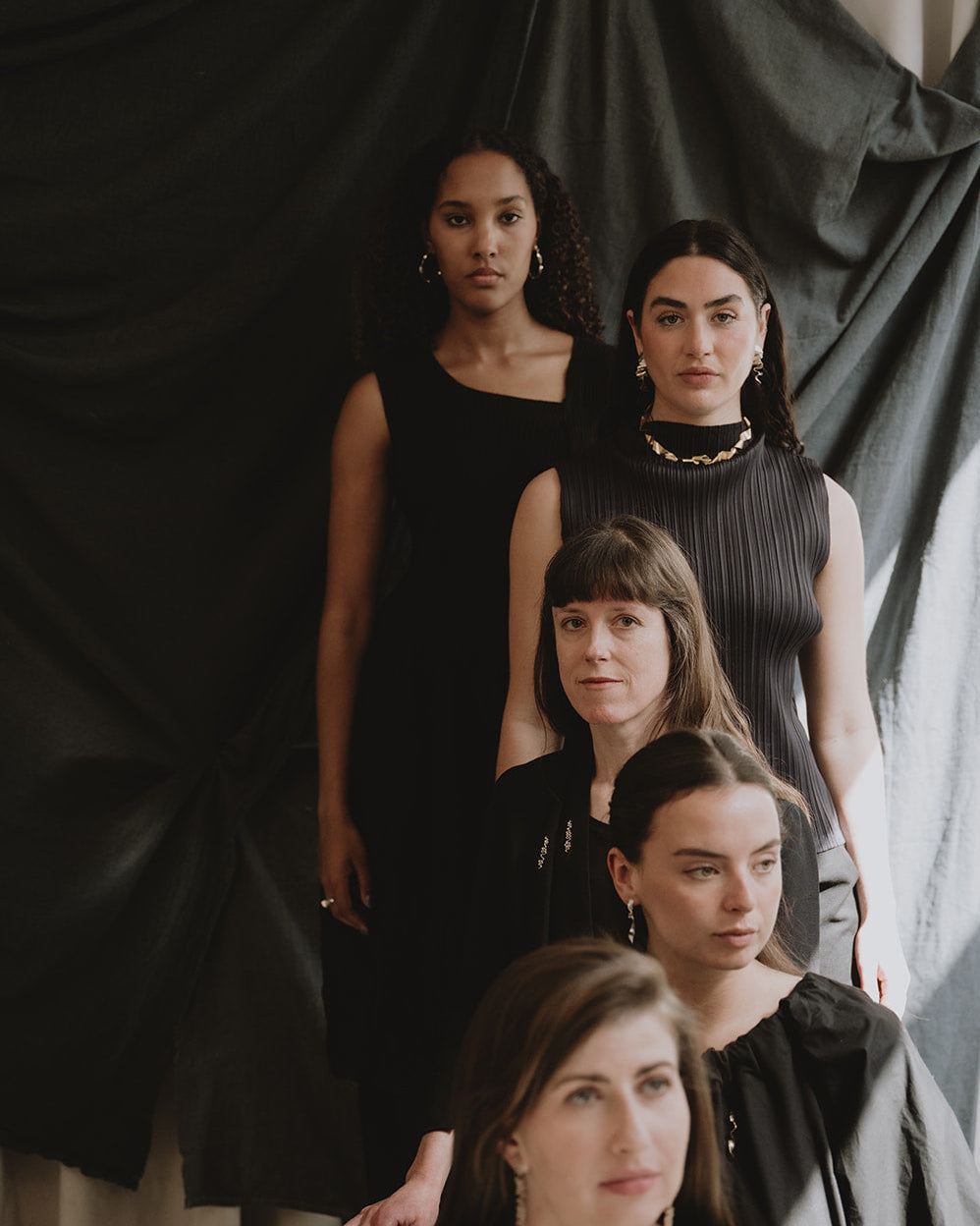 Five women posing together in black clothing against a dark draped fabric backdrop in an indoor studio setting.