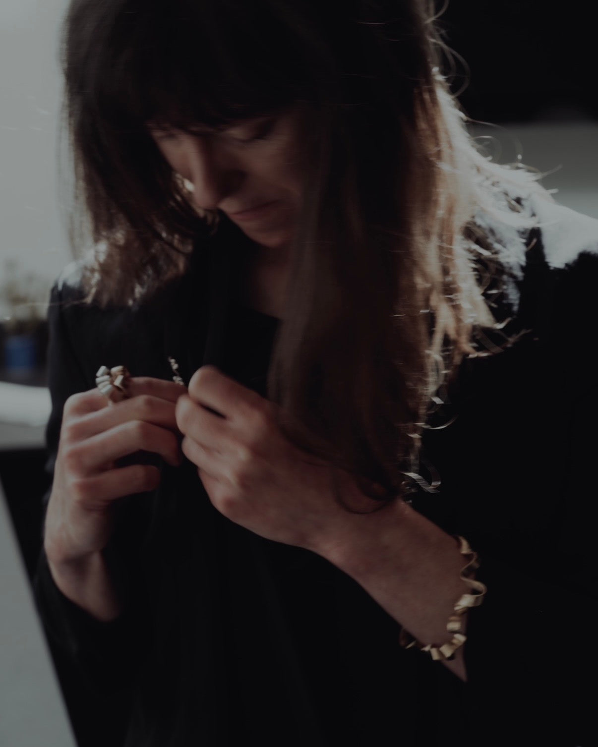 A woman with long dark hair looks down while holding small jewelry pieces in her hands. She is wearing a black top and a gold beaded bracelet, photographed in a dim indoor setting.
