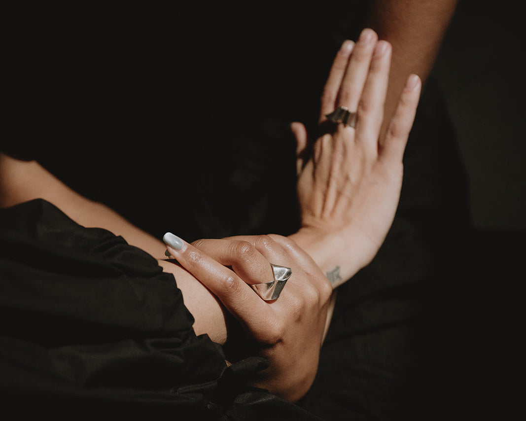 A close-up lifestyle shot of a person's hands and arms, showcasing a silver geometric ring on one finger and a dark band ring on another, set against a dark black background.