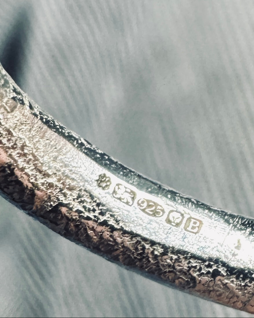 Close-up of a textured silver-toned metal band with engraved hallmarks including '925' and a heart symbol, photographed against a gray brushed background.