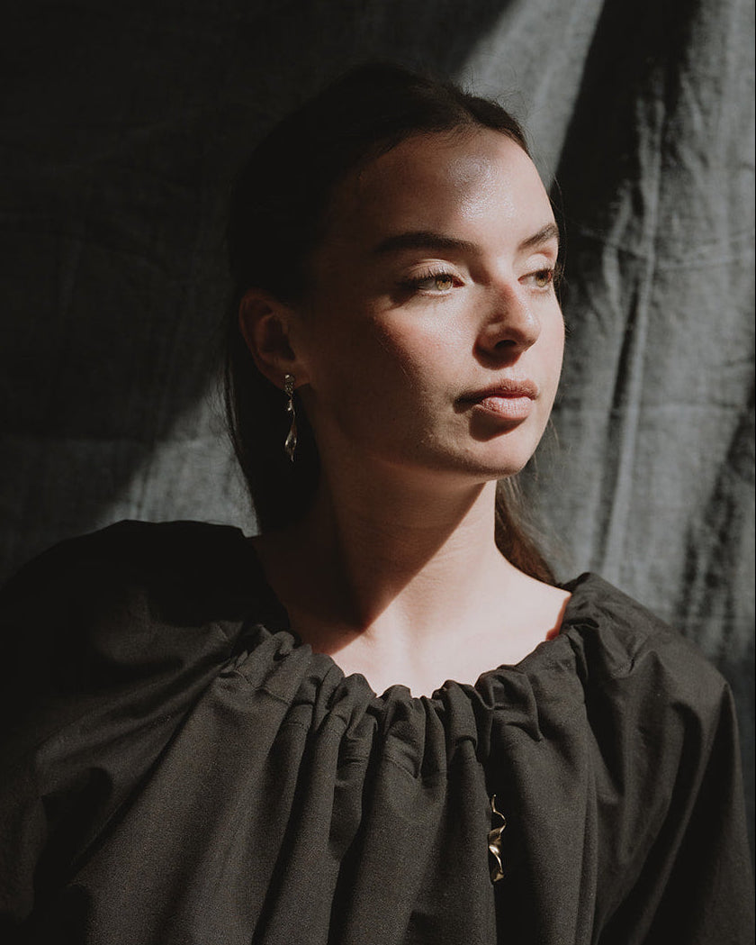 A woman wearing a dark, loose-fitting blouse with dramatic side-slit lighting against a dark draped backdrop.