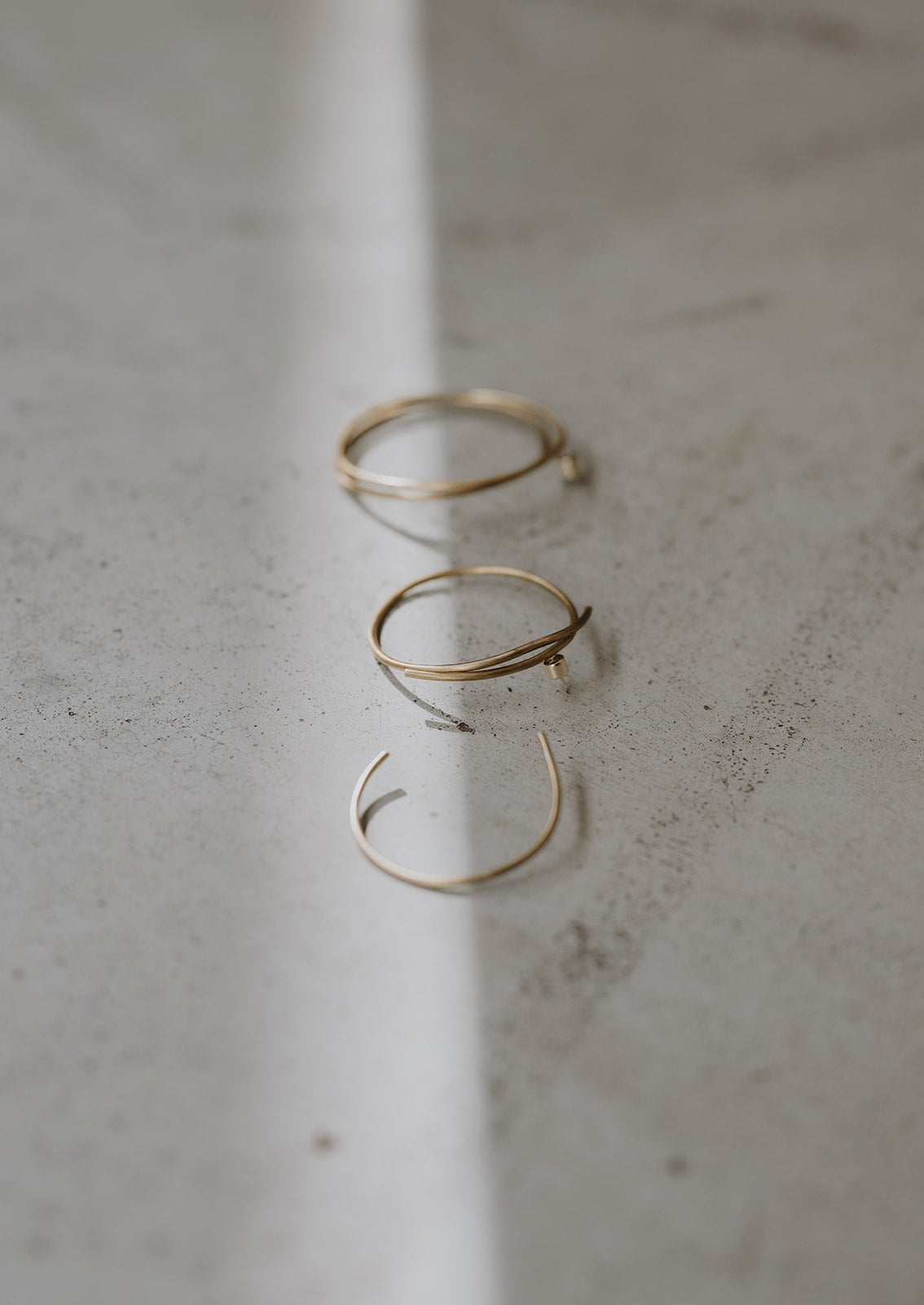 Brass wire hoop earrings, three in a row lay on a concrete floor with light streaming over the pieces.