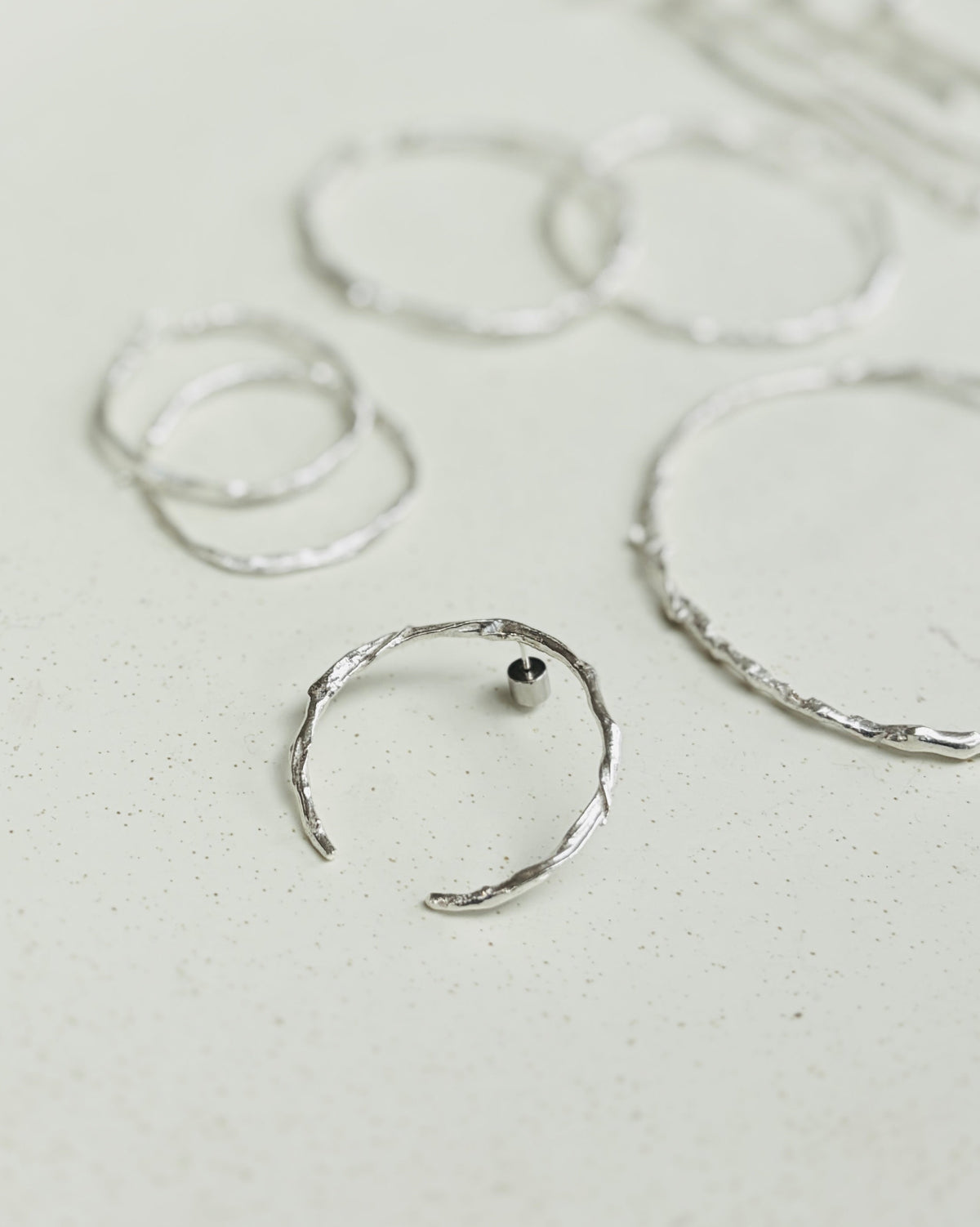 A set of textured silver hoop and ring earrings arranged on a light beige surface, with some pieces in the background out of focus.