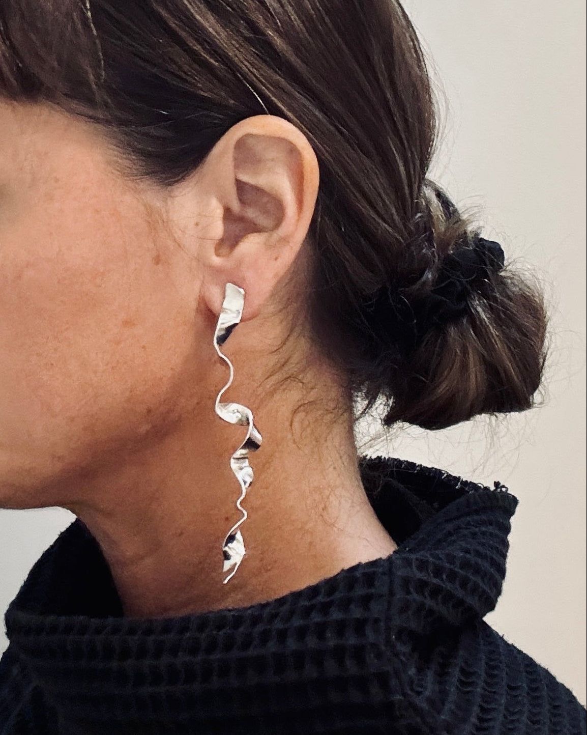 A woman in profile wearing a long, silver, spiral-shaped dangle earring and a black textured waffle-knit turtleneck sweater.