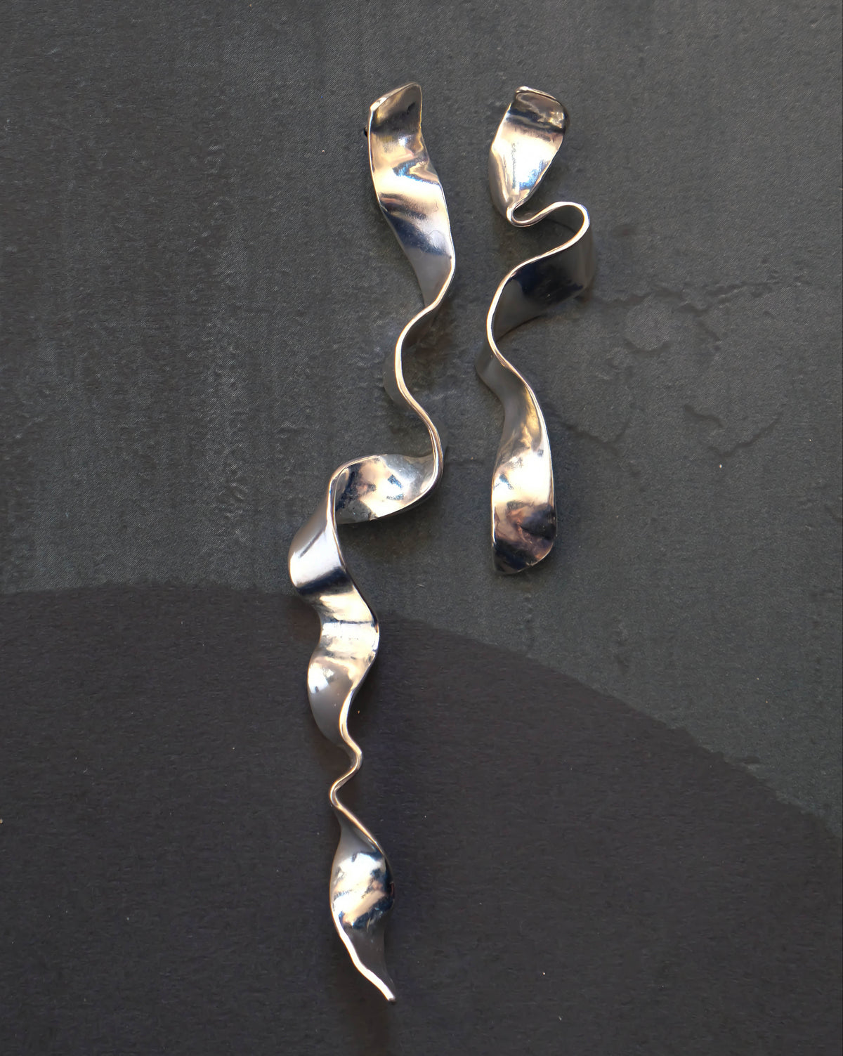 Two metallic silver wavy ribbon-like accessories laid flat on a dark charcoal background.