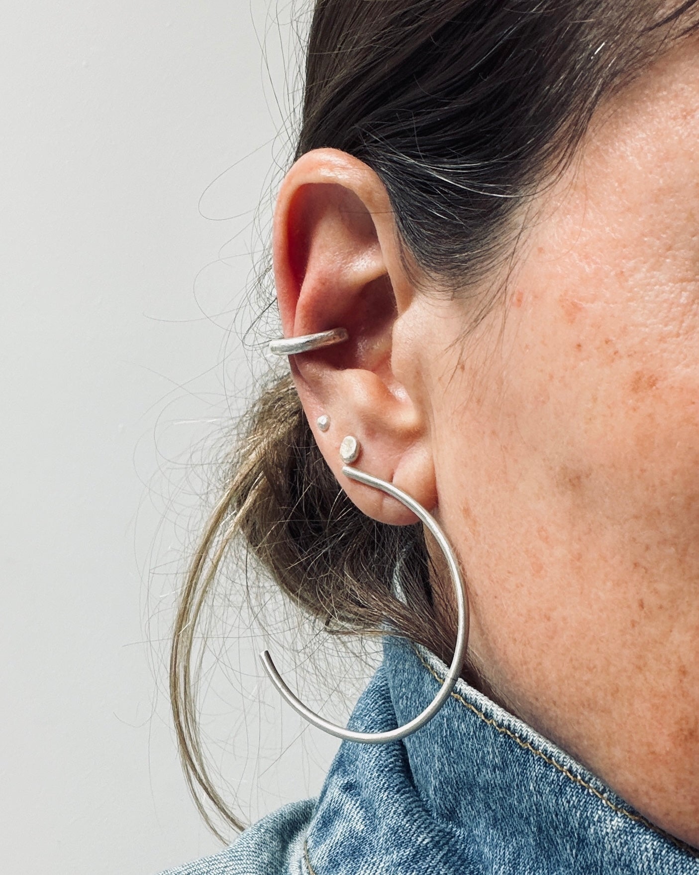 A close-up of a person's ear wearing multiple silver earrings, including a large curved hoop earring, a smaller curved hoop, and small stud earrings, against a light gray background.